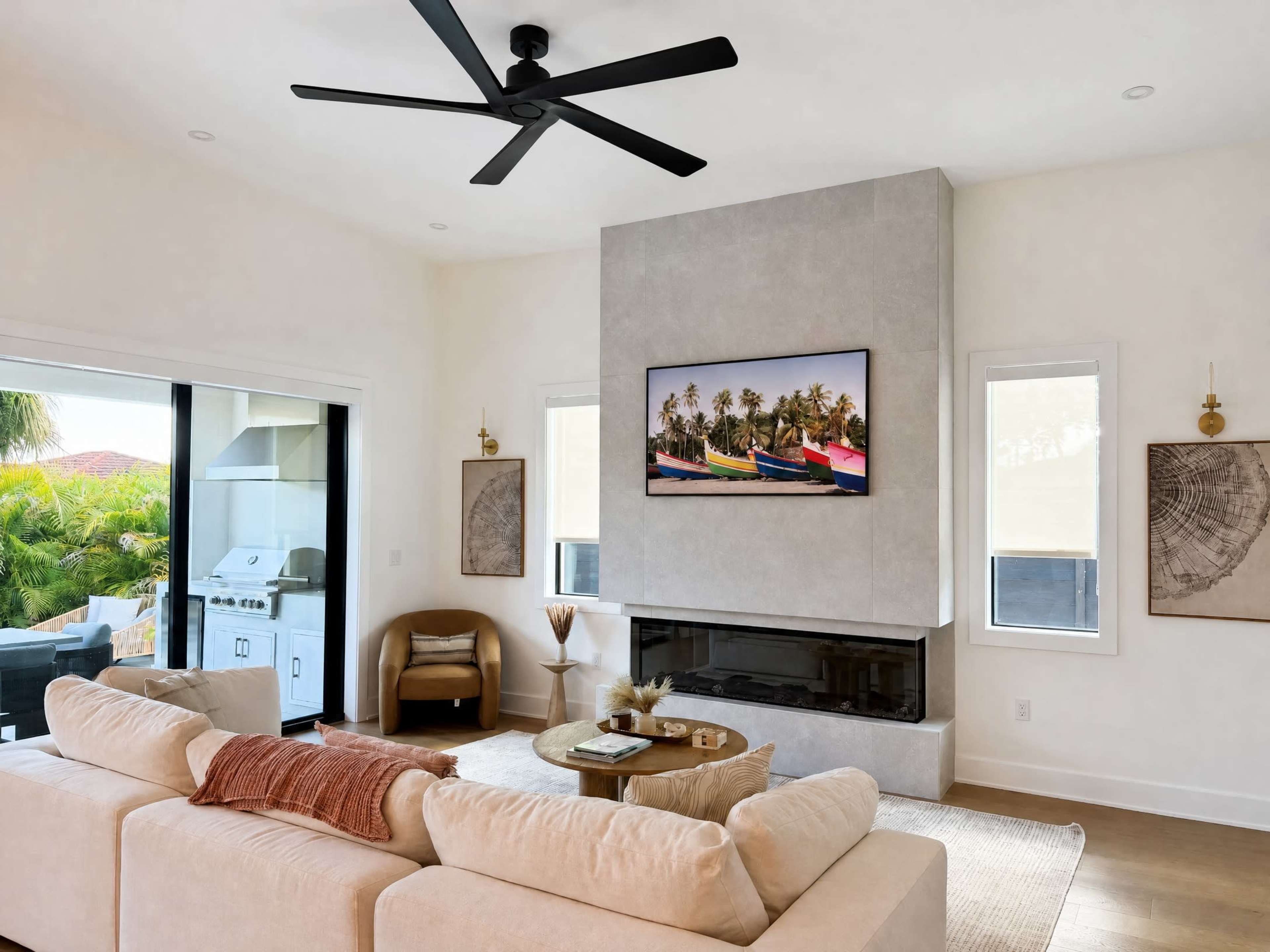 The image shows a modern living room with a beige sectional sofa facing a wall-mounted television above a minimalist fireplace, flanked by large windows and a sliding door leading to an outdoor area.