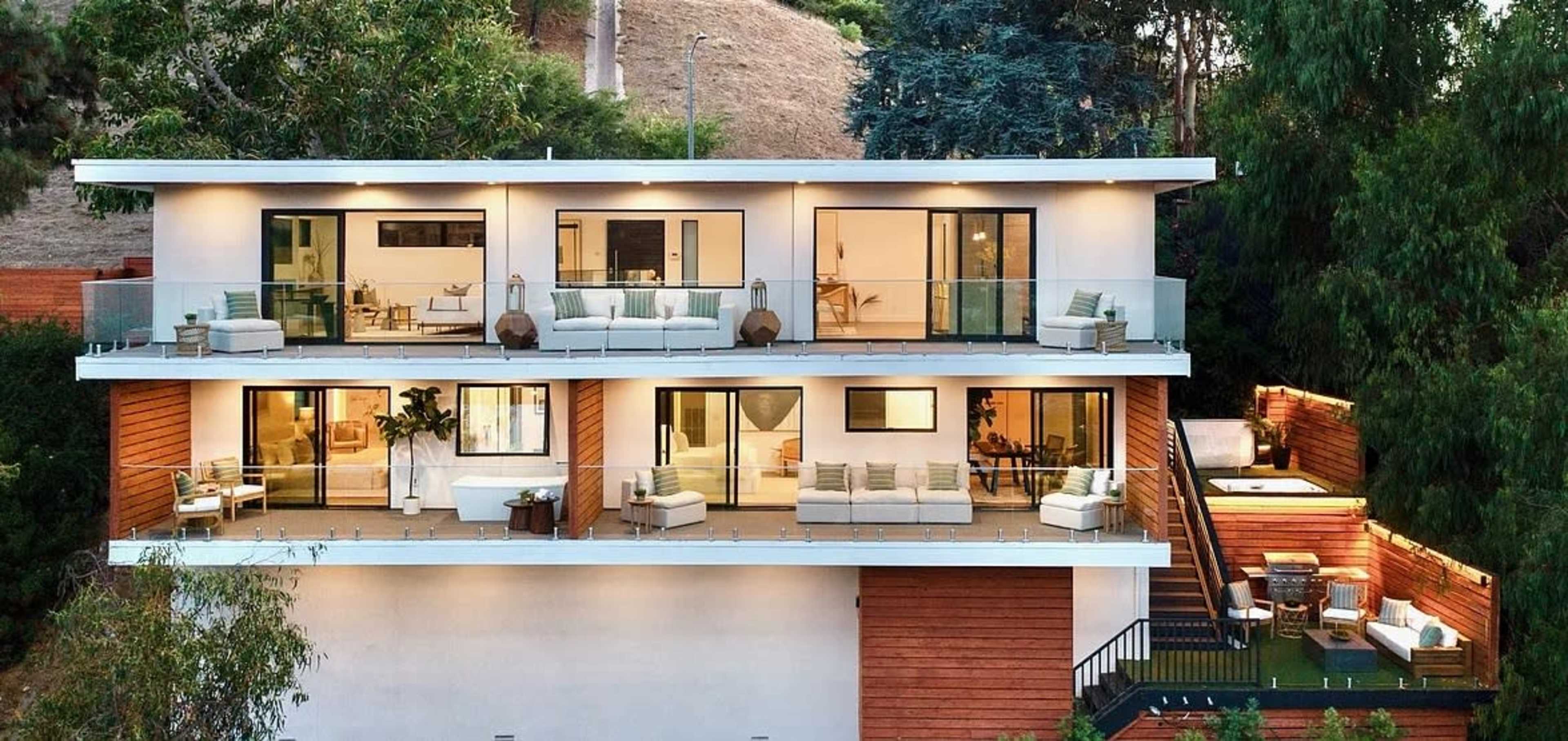The image shows a modern, multi-level house with large glass doors and balconies, surrounded by greenery.