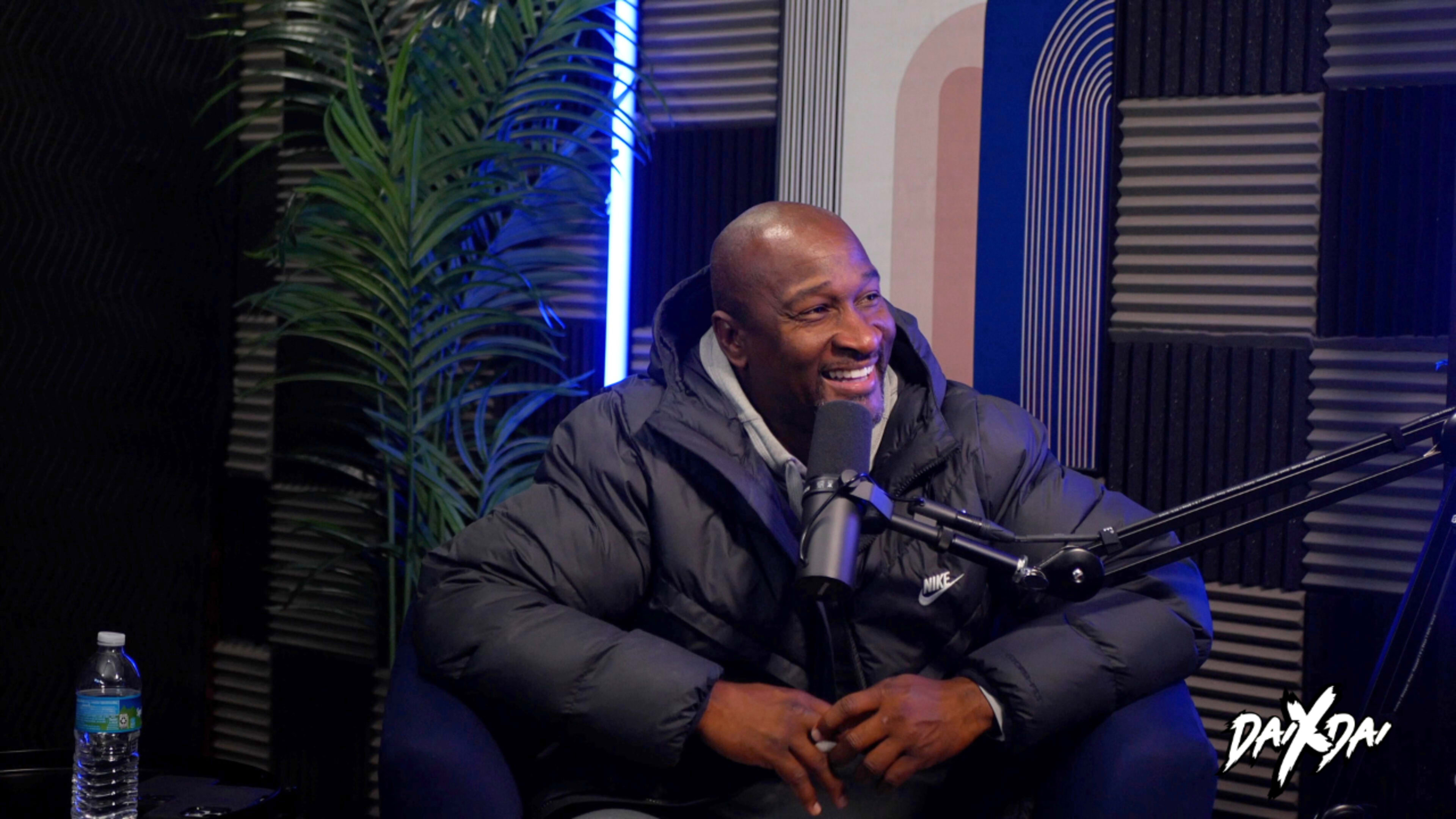 A man in a black jacket sits in a recording studio with a microphone, surrounded by soundproofing panels and a green plant.