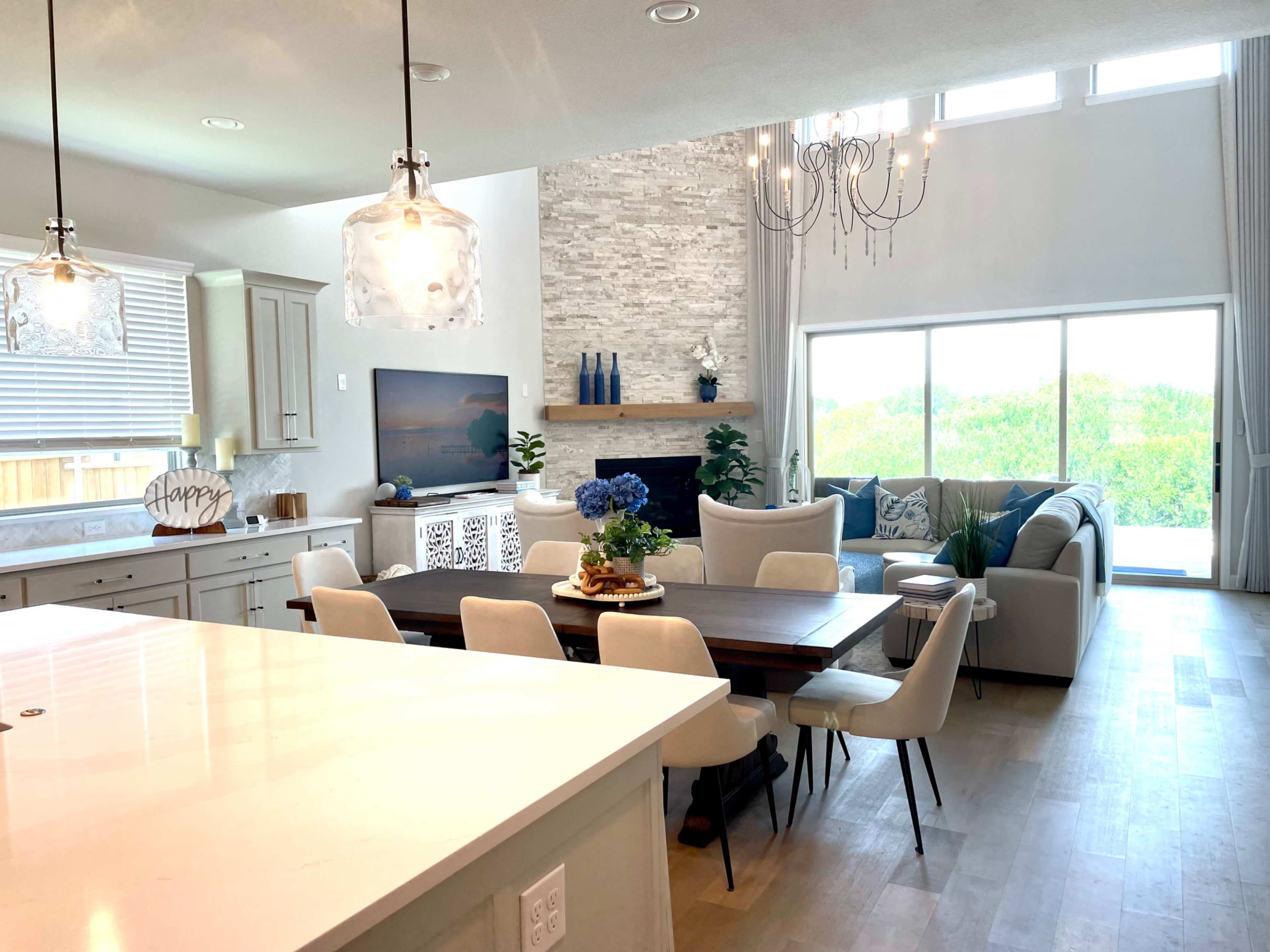 A modern open-concept living space with a dining area, a kitchen with a large island, and a lounge featuring a stone fireplace and large windows.