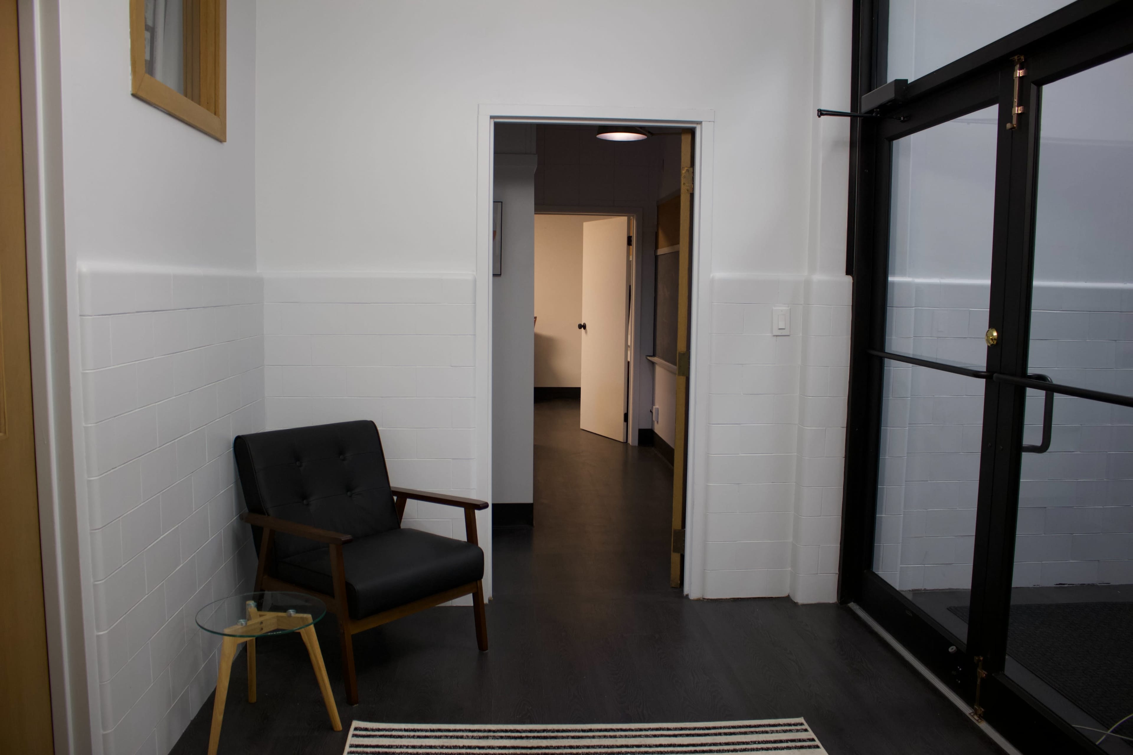 A minimalist entryway featuring a black chair next to a small table, with a door leading to a hallway in the background.