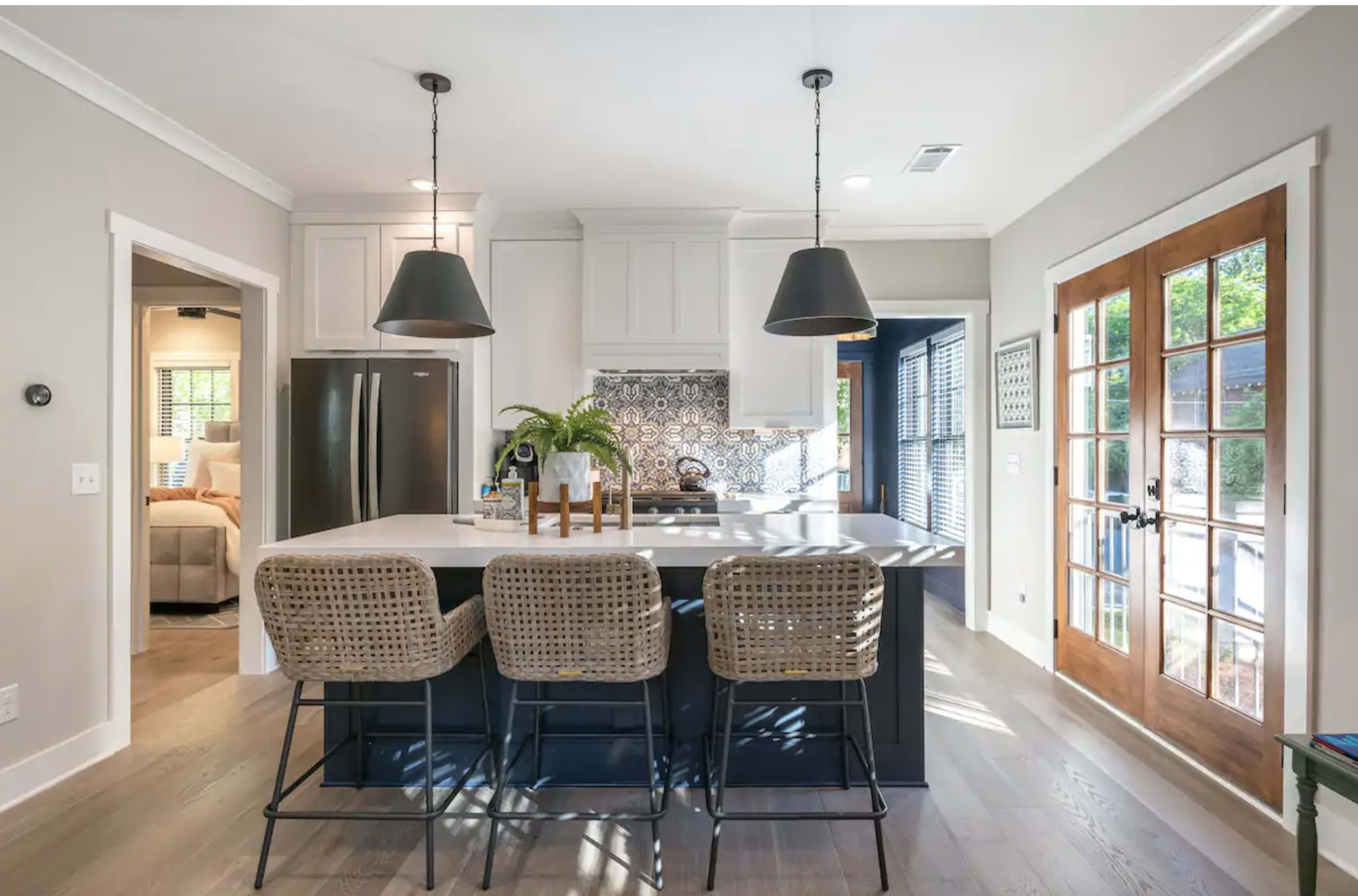 The image shows a modern kitchen with a central island featuring three wicker bar stools, pendant lights above, and a patterned backsplash, adjacent to a doorway leading to a living area.