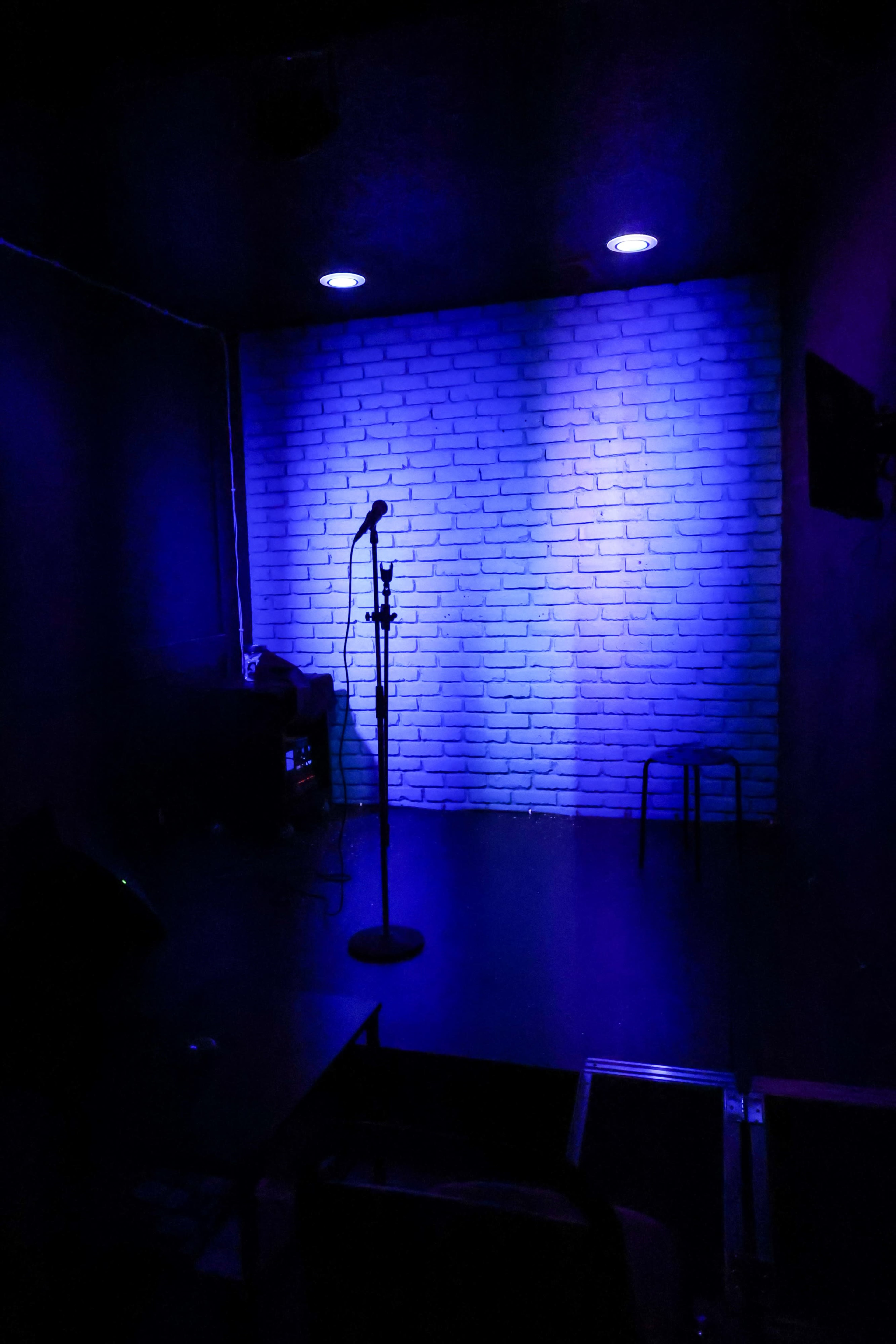The image shows a dimly lit performance space with a microphone on a stand in front of a white brick wall, illuminated by blue lights.