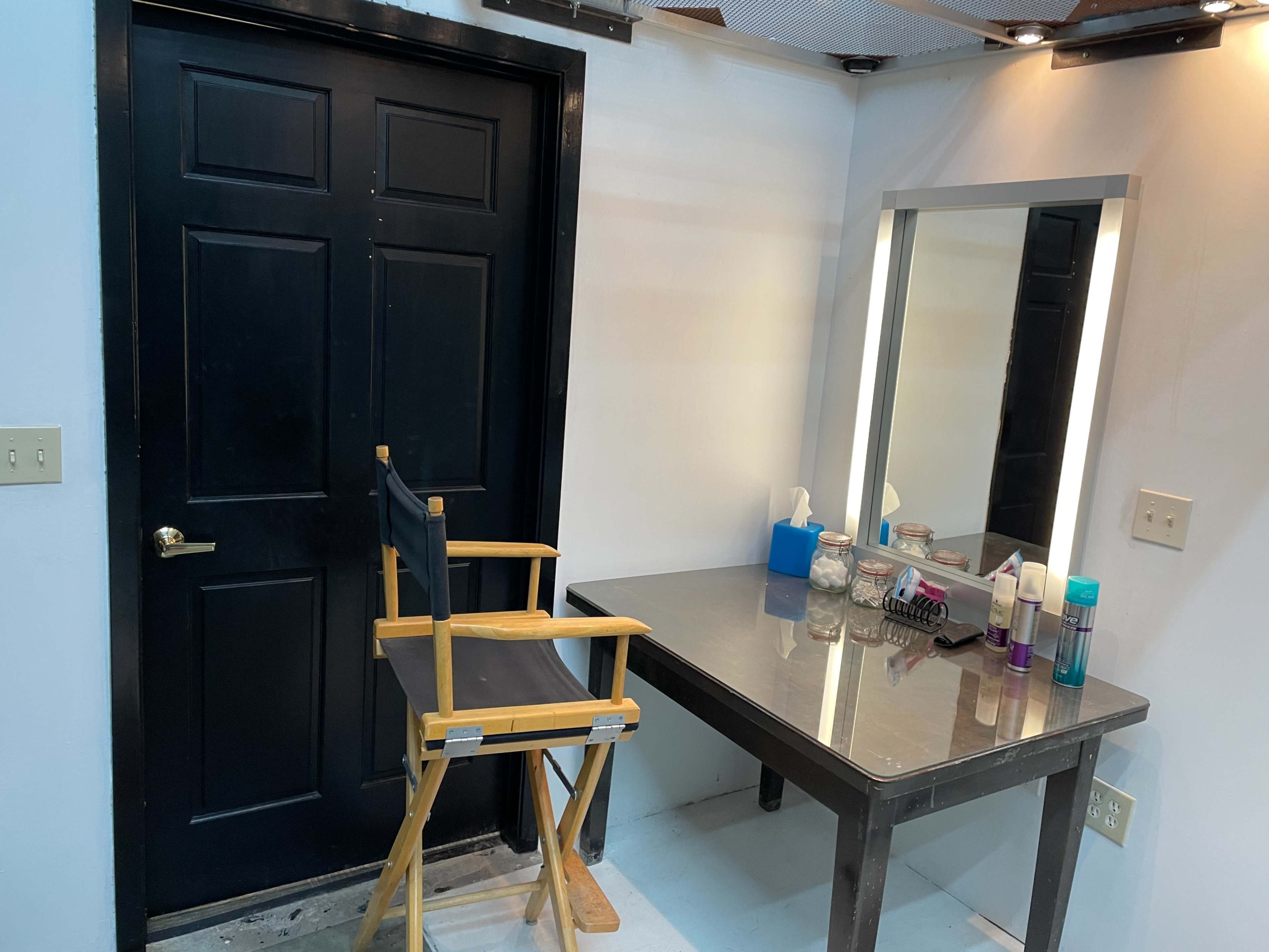 A makeup room with a black door, a wooden director's chair, a mirror with lights, and a table equipped with various cosmetic products.