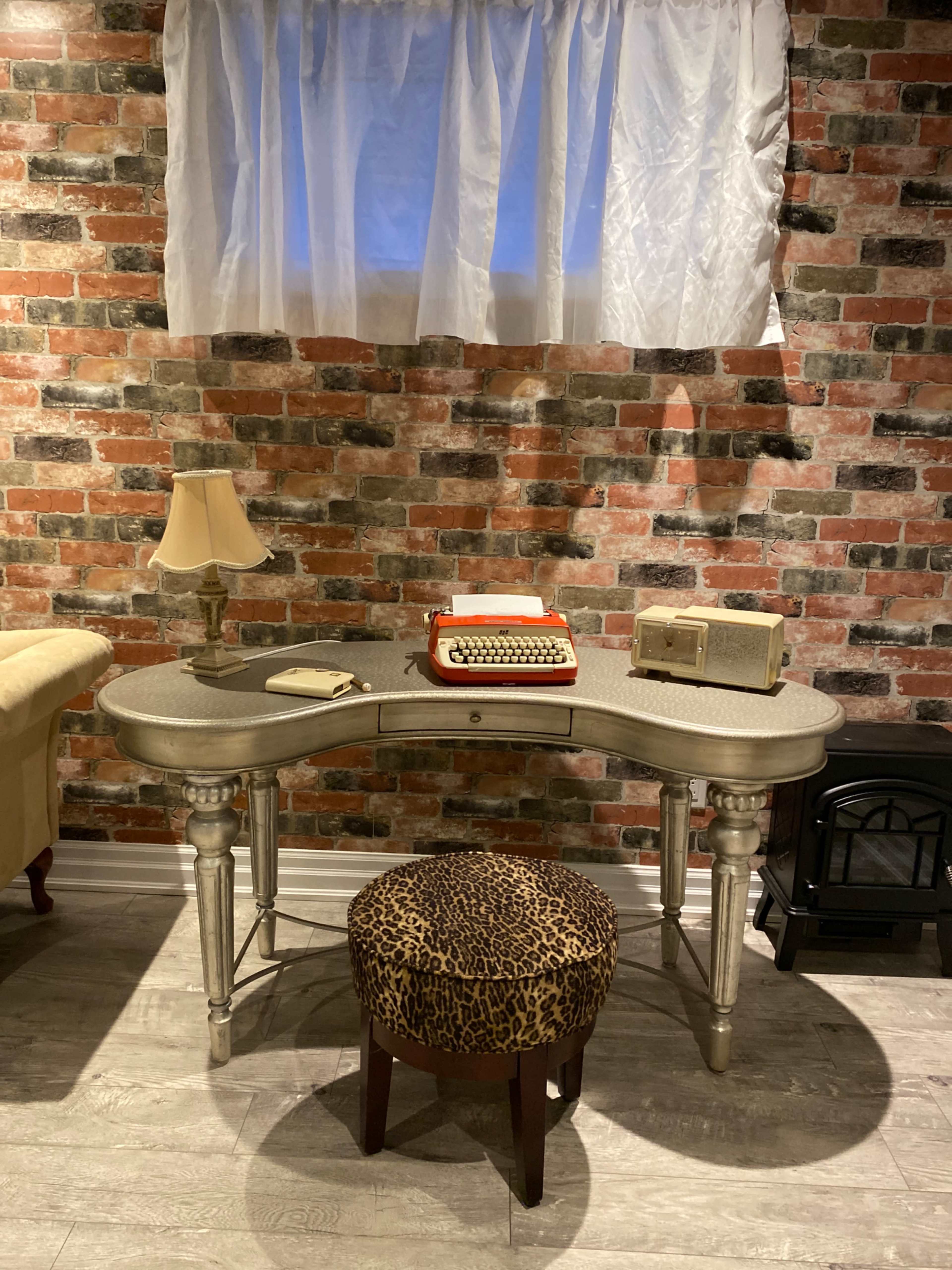 A vintage-style desk with a typewriter and a decorative box sits against a brick wall, accompanied by a lamp and a stool with a leopard print pattern.