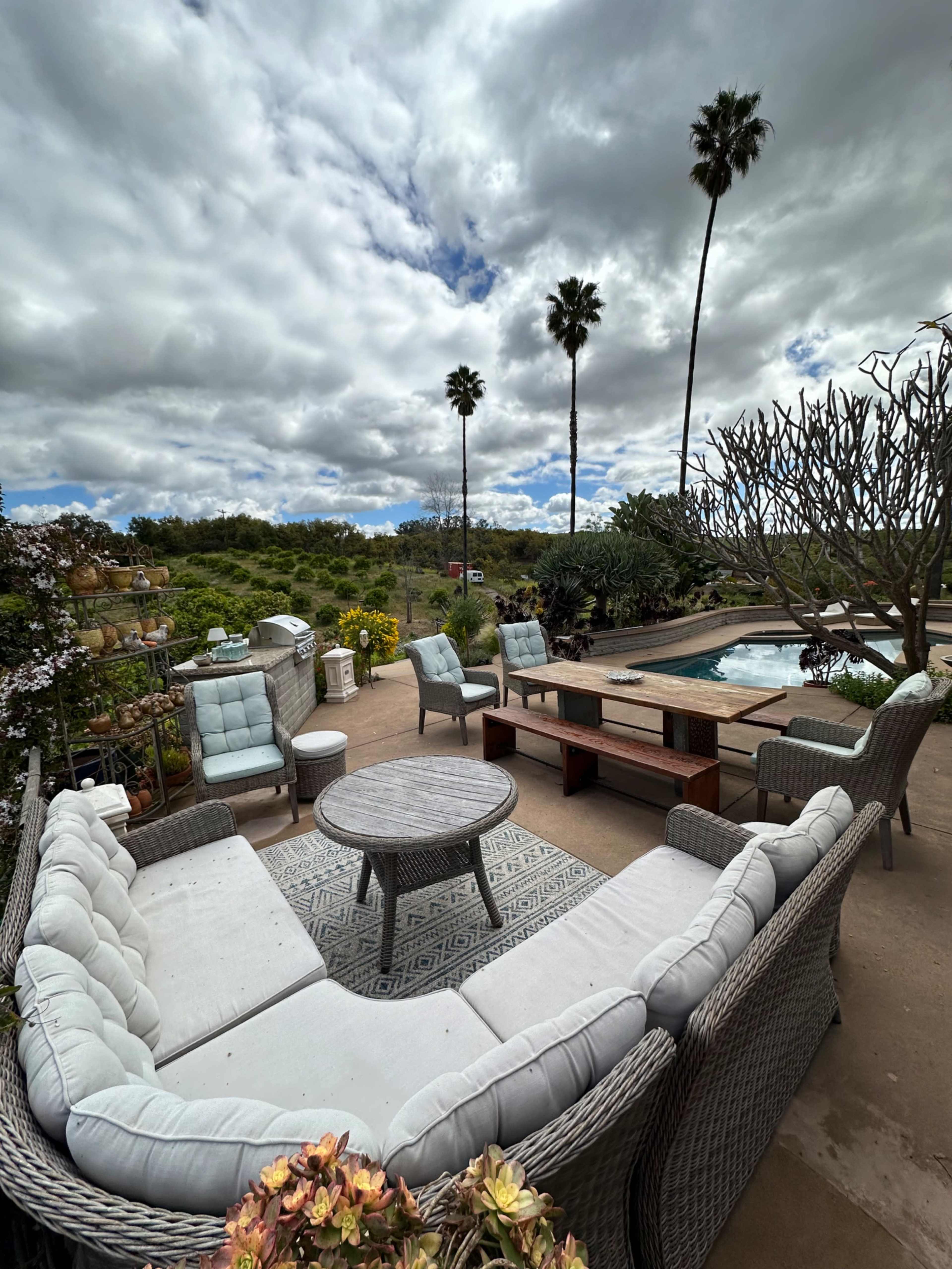 A patio area featuring a circular seating arrangement with a central table, surrounded by greenery and a pool under a cloudy sky.
