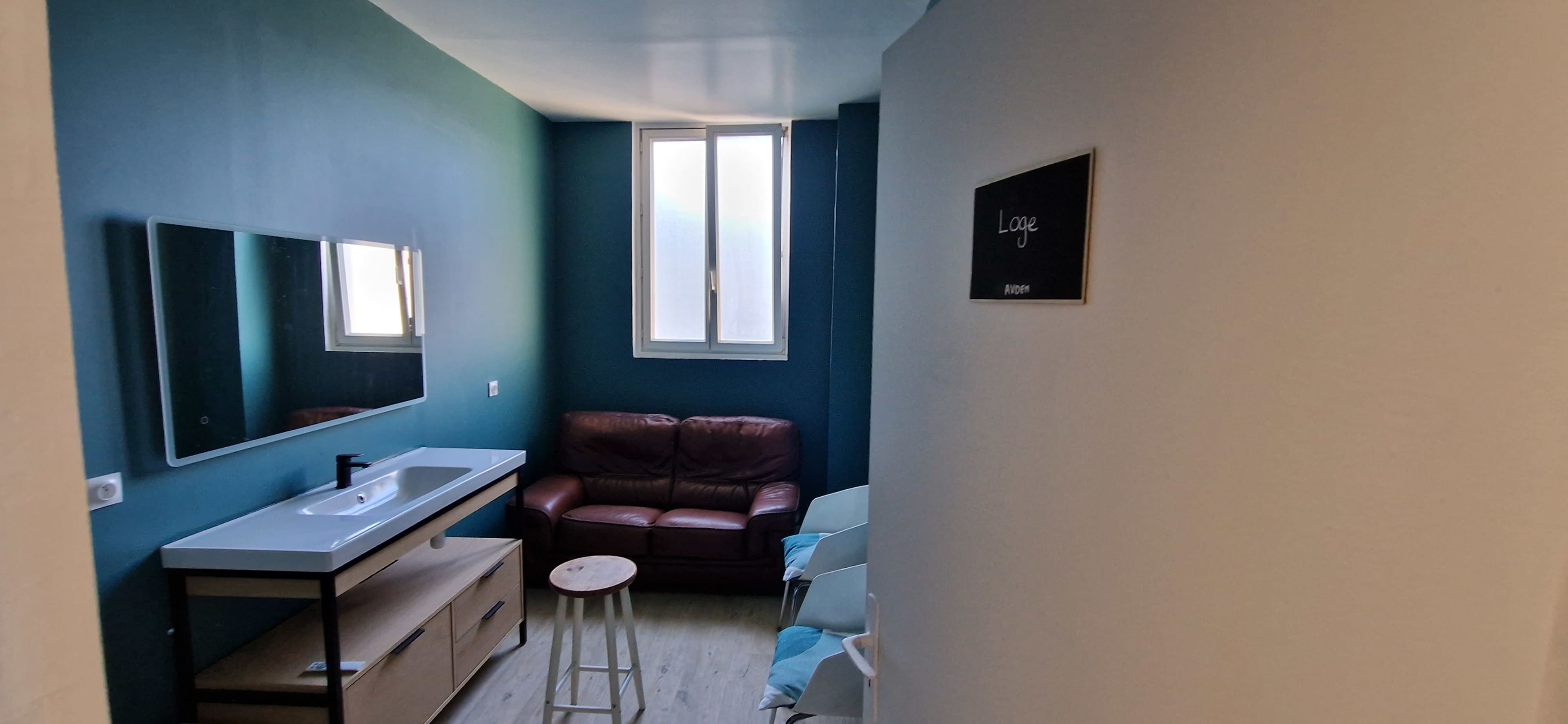 A small room with a brown leather couch, a sink with a mirror above it, and a window letting in light.