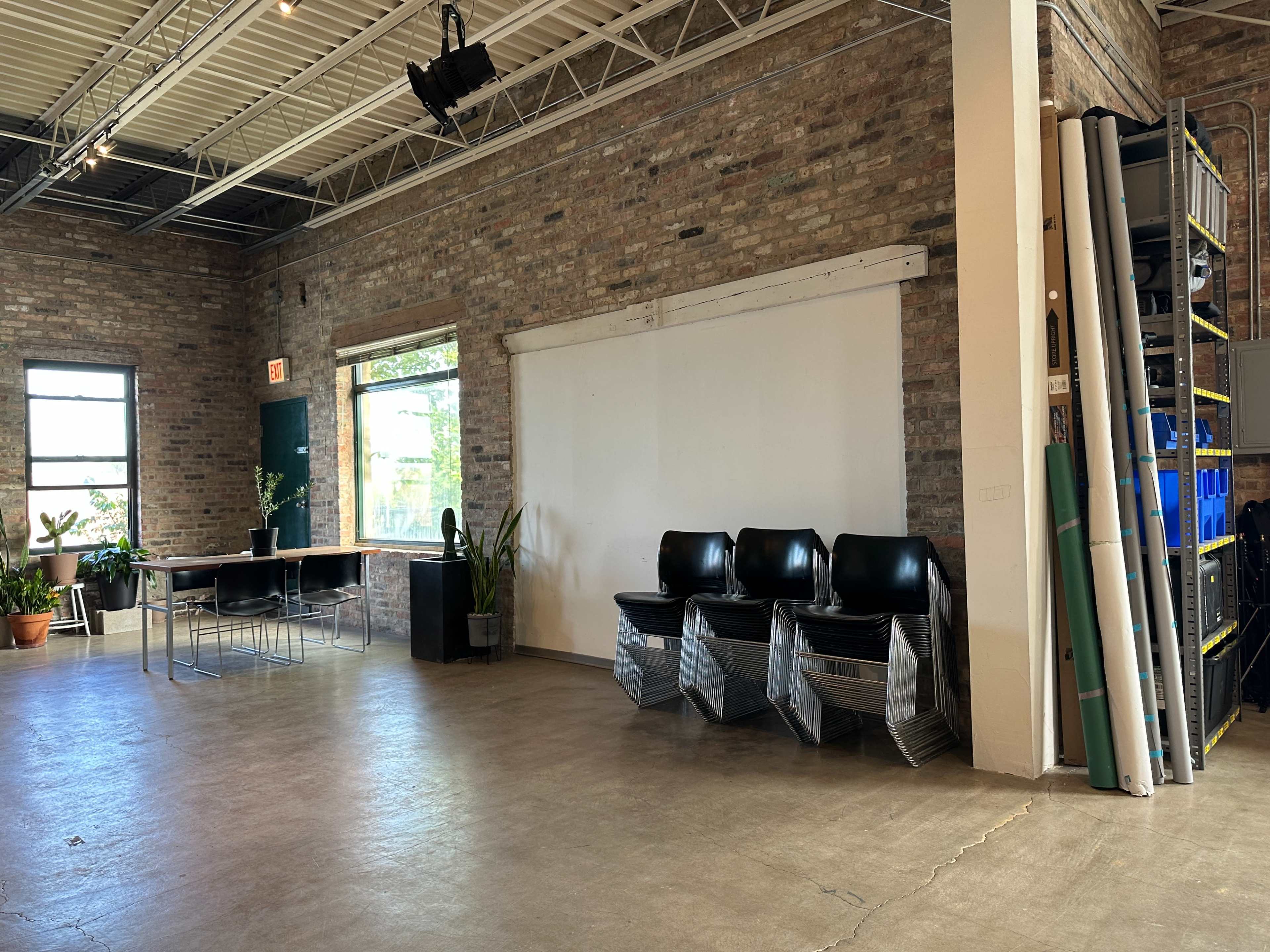 The image shows a spacious room with brick walls, featuring a table and chairs on one side and stacked chairs near a blank white wall, with shelves of stacked containers and rolled materials on the other side.