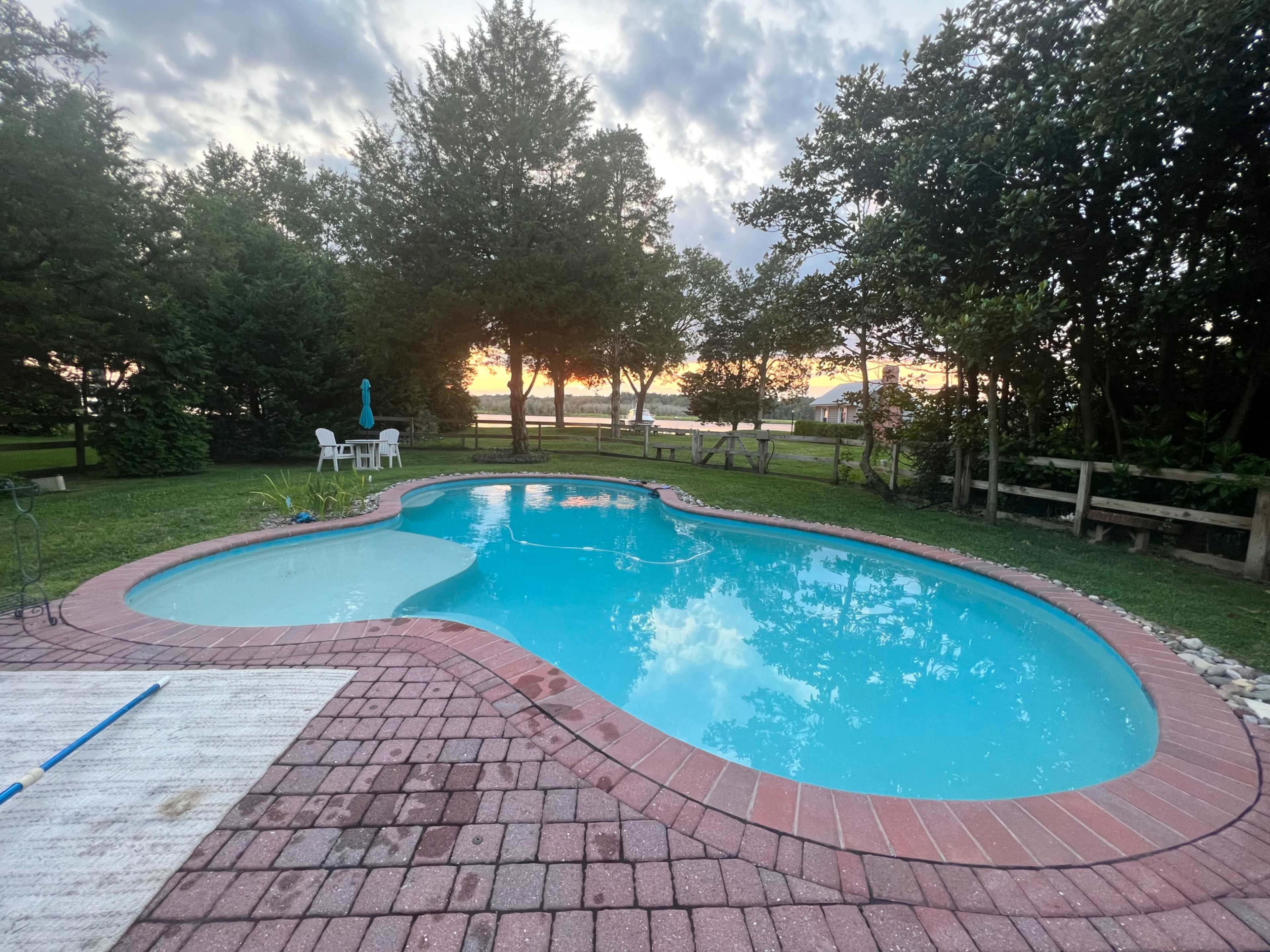 A swimming pool with a brick patio surrounded by greenery, set against a sunset backdrop.