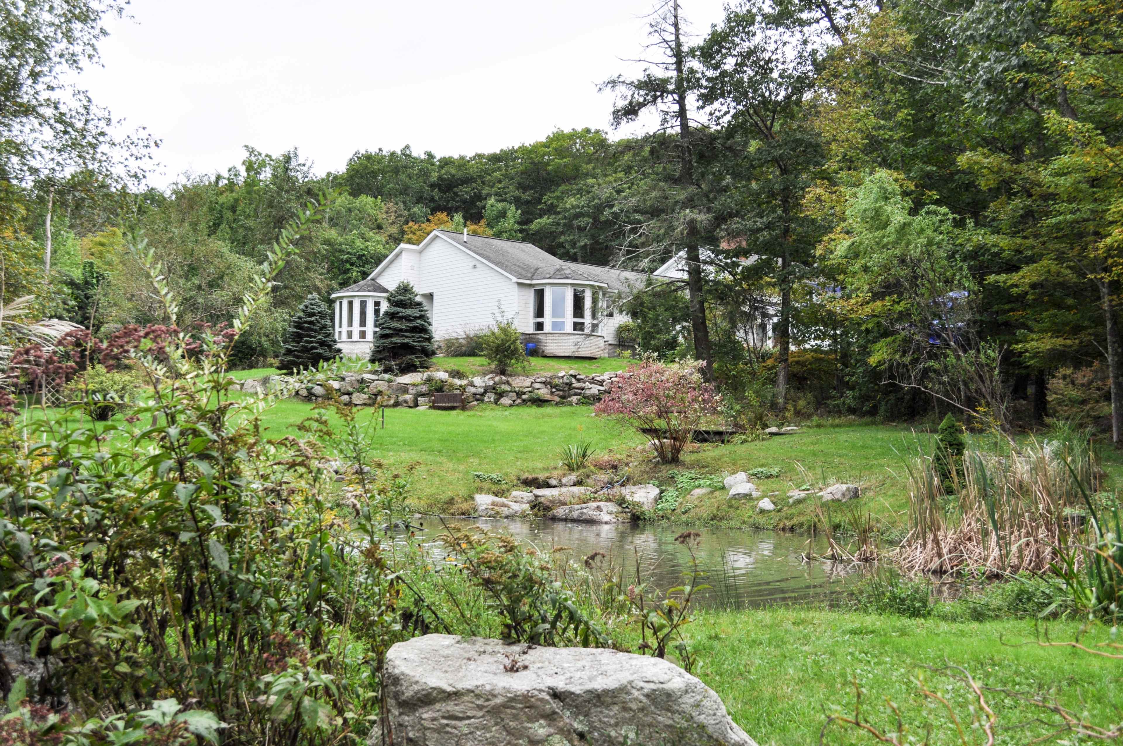 A white house is situated near a pond, surrounded by lush greenery and landscaping in a wooded area.
