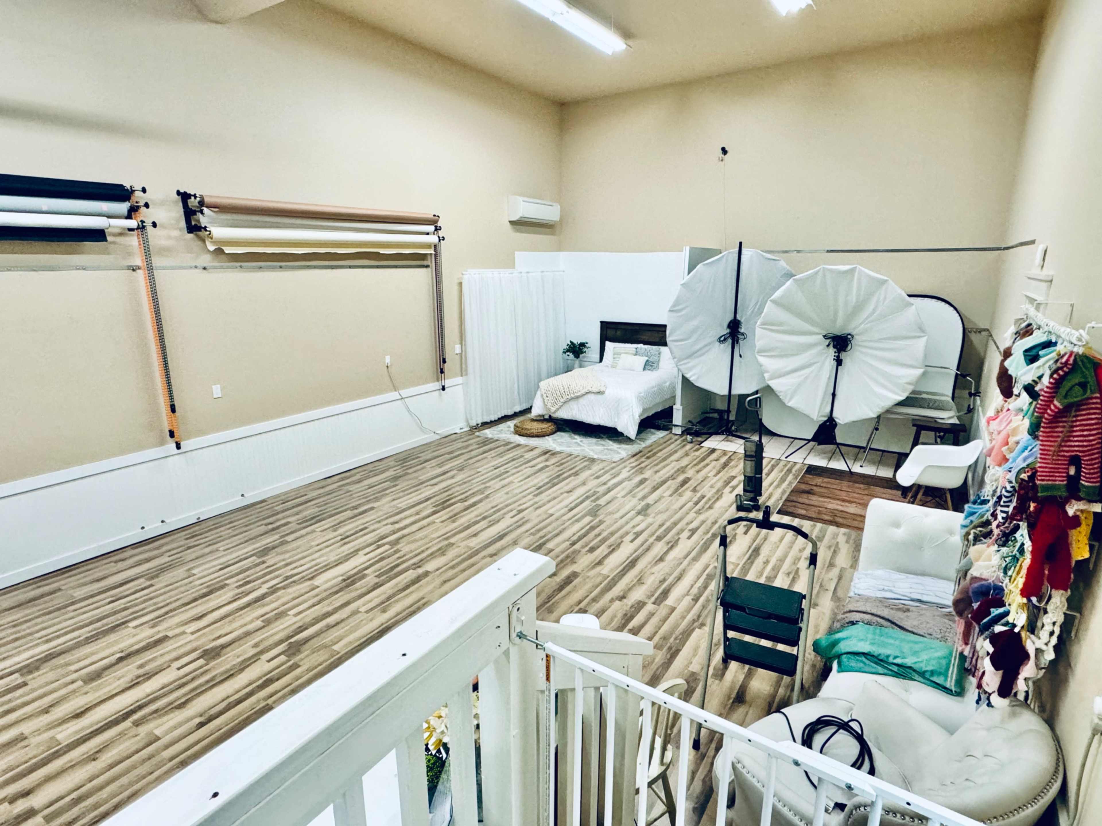 The image shows a spacious studio with a wooden floor, featuring a bed, photography backdrop rolls, large lighting equipment, and a seating area.