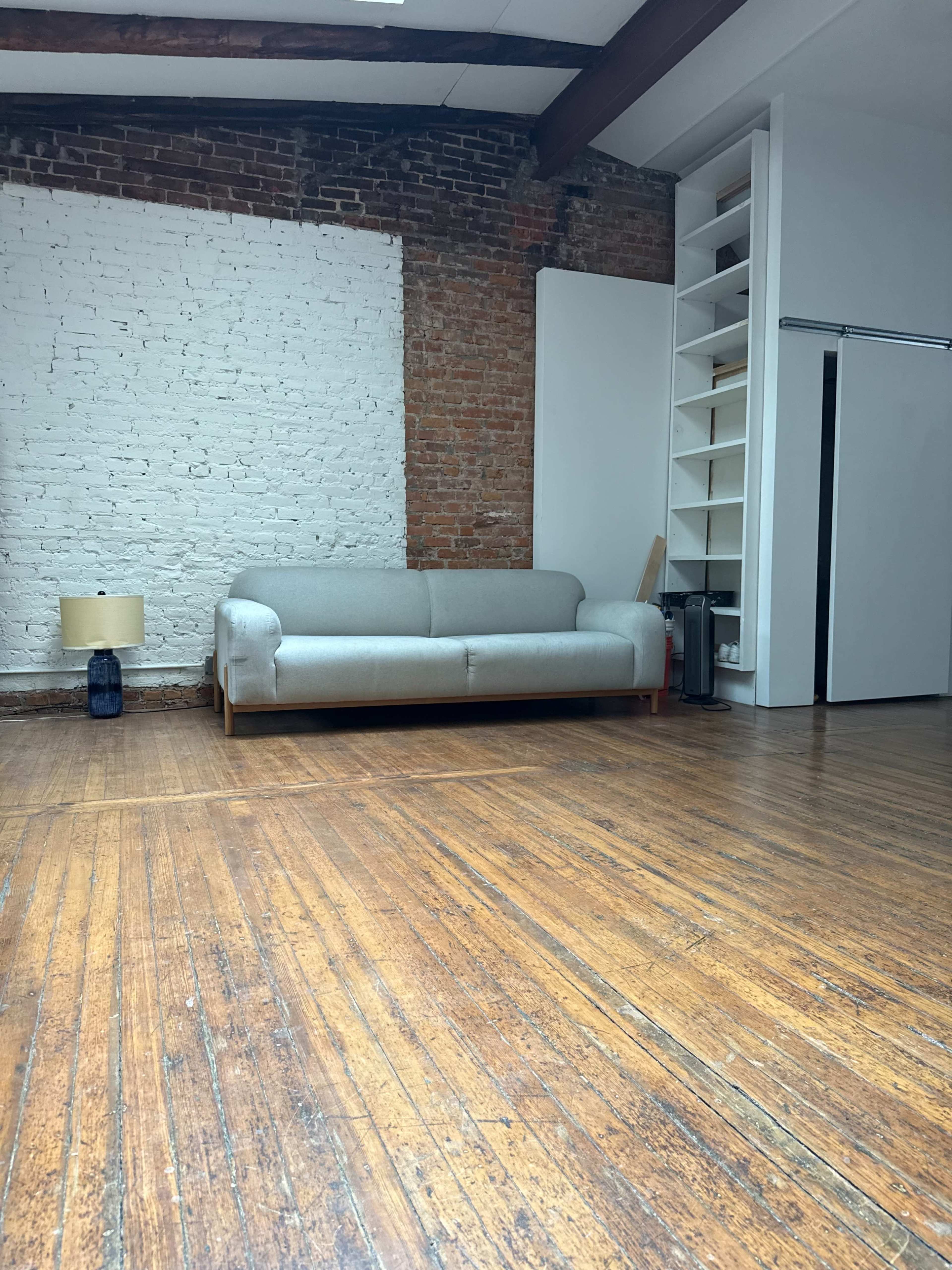 The image shows a simple living space featuring a light-colored sofa against a brick wall, wooden flooring, and a lamp on the floor.