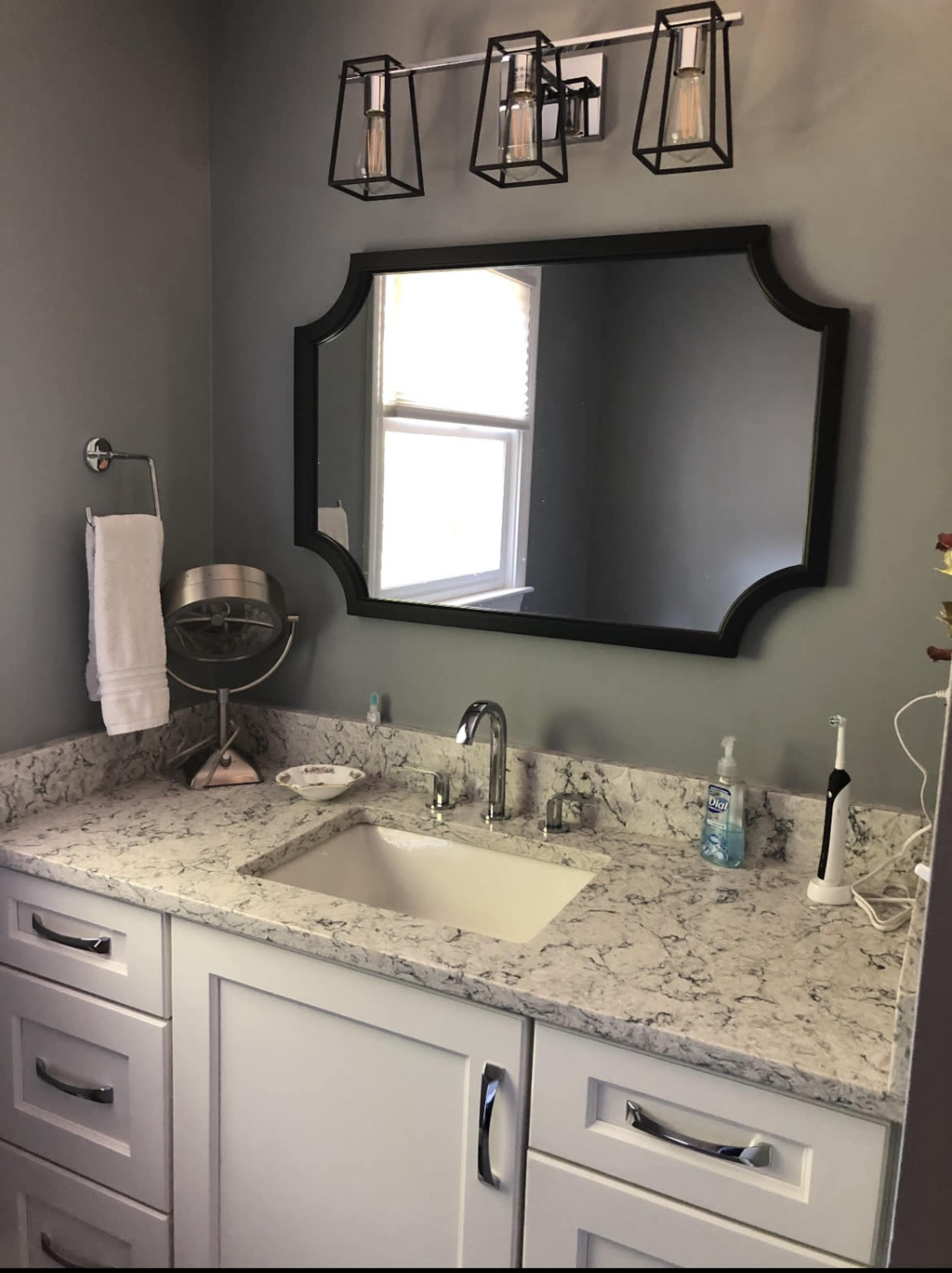 A bathroom features a white sink and countertop with a large black-framed mirror above, complemented by a modern light fixture and various bathroom accessories.
