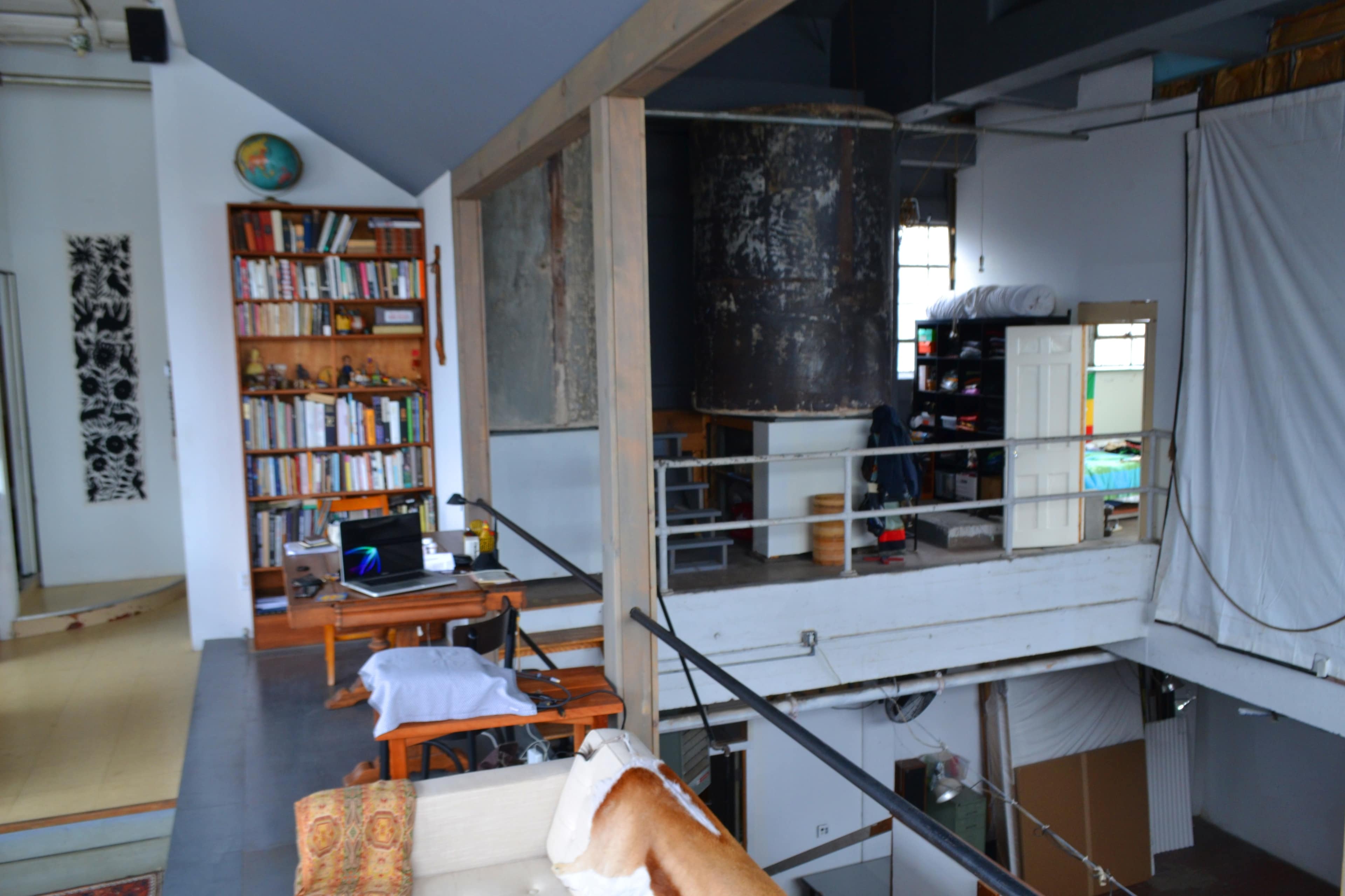 A spacious, modern loft with a split-level layout, featuring a bookshelf, a desk with a computer, and a partially visible kitchen area.
