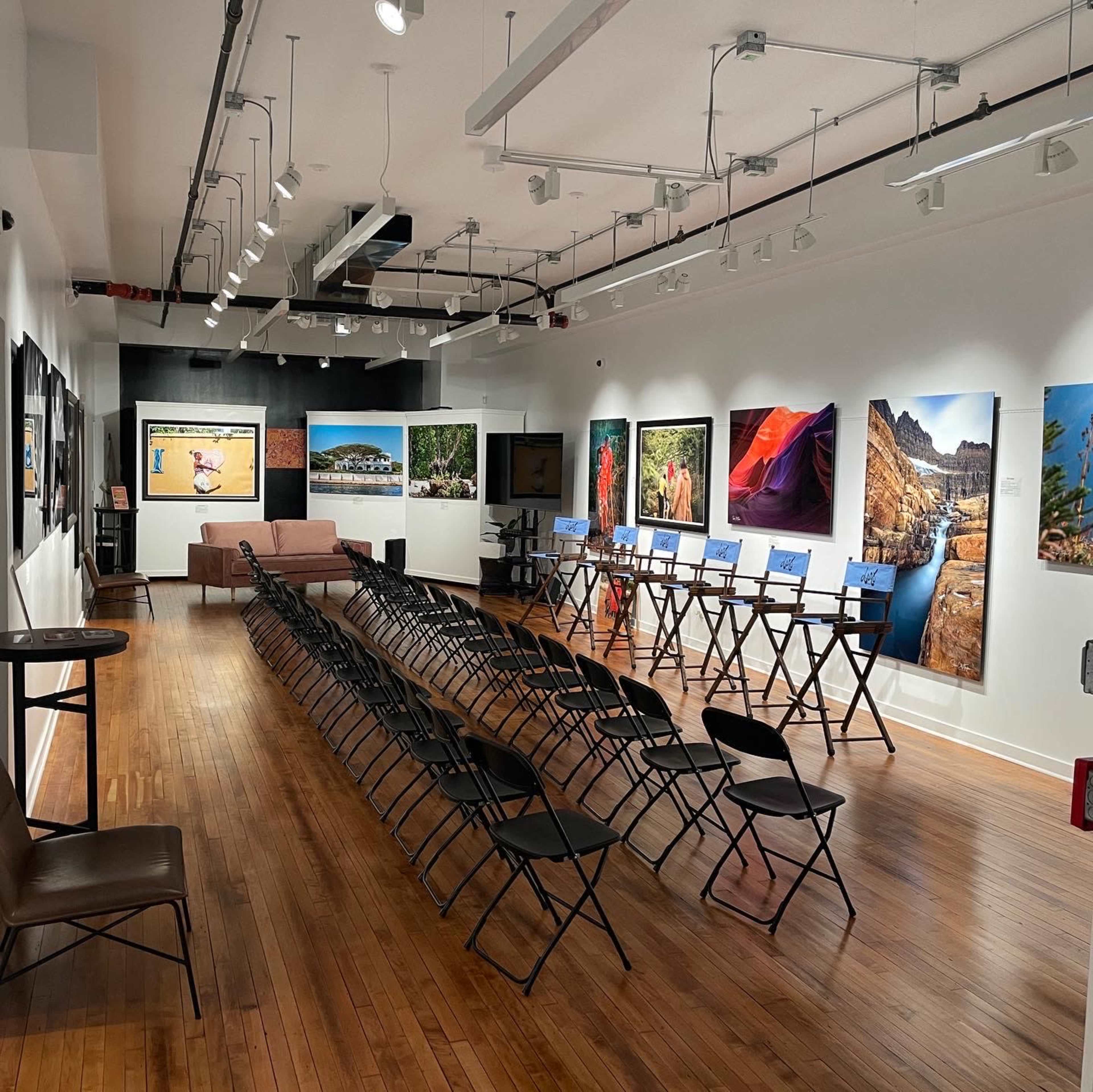 An art gallery with wooden floors, rows of folding chairs facing large photographs displayed on the walls, and a couch in the corner.