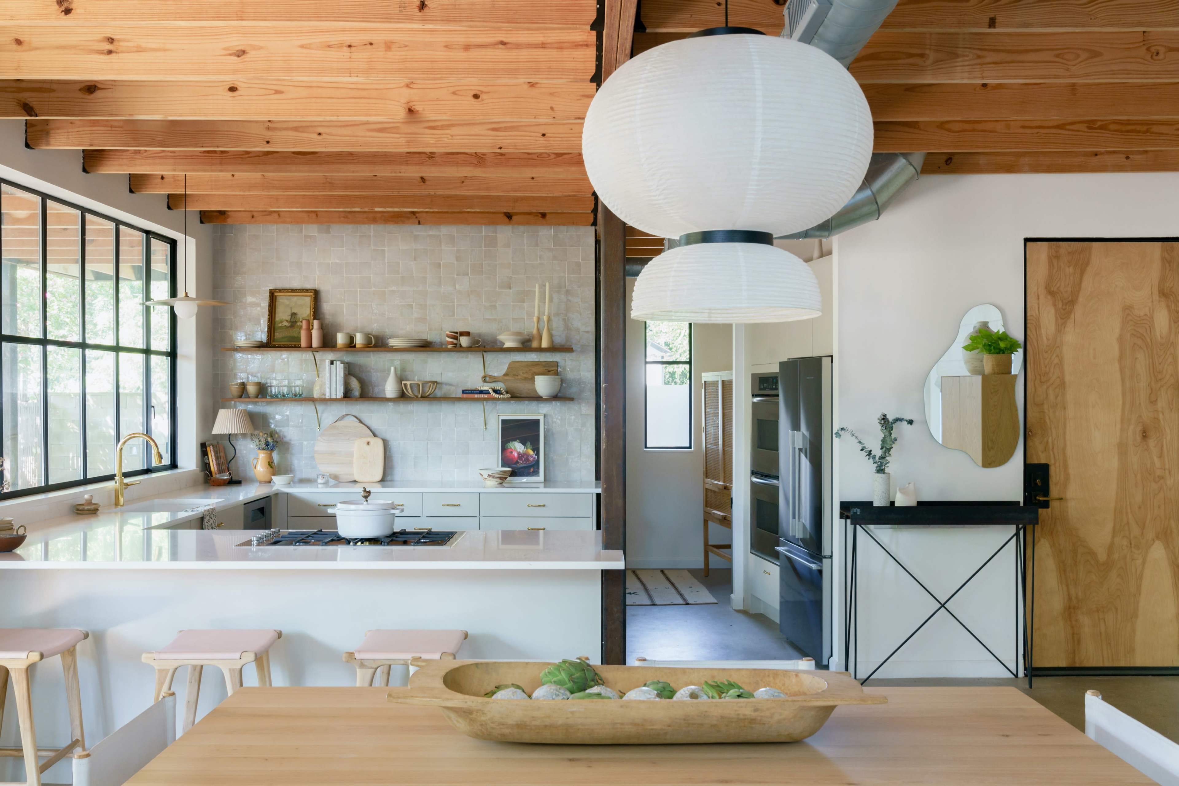 A modern kitchen features a central island with minimalistic decor, wooden beams overhead, and a light fixture hanging above a wooden dining table.