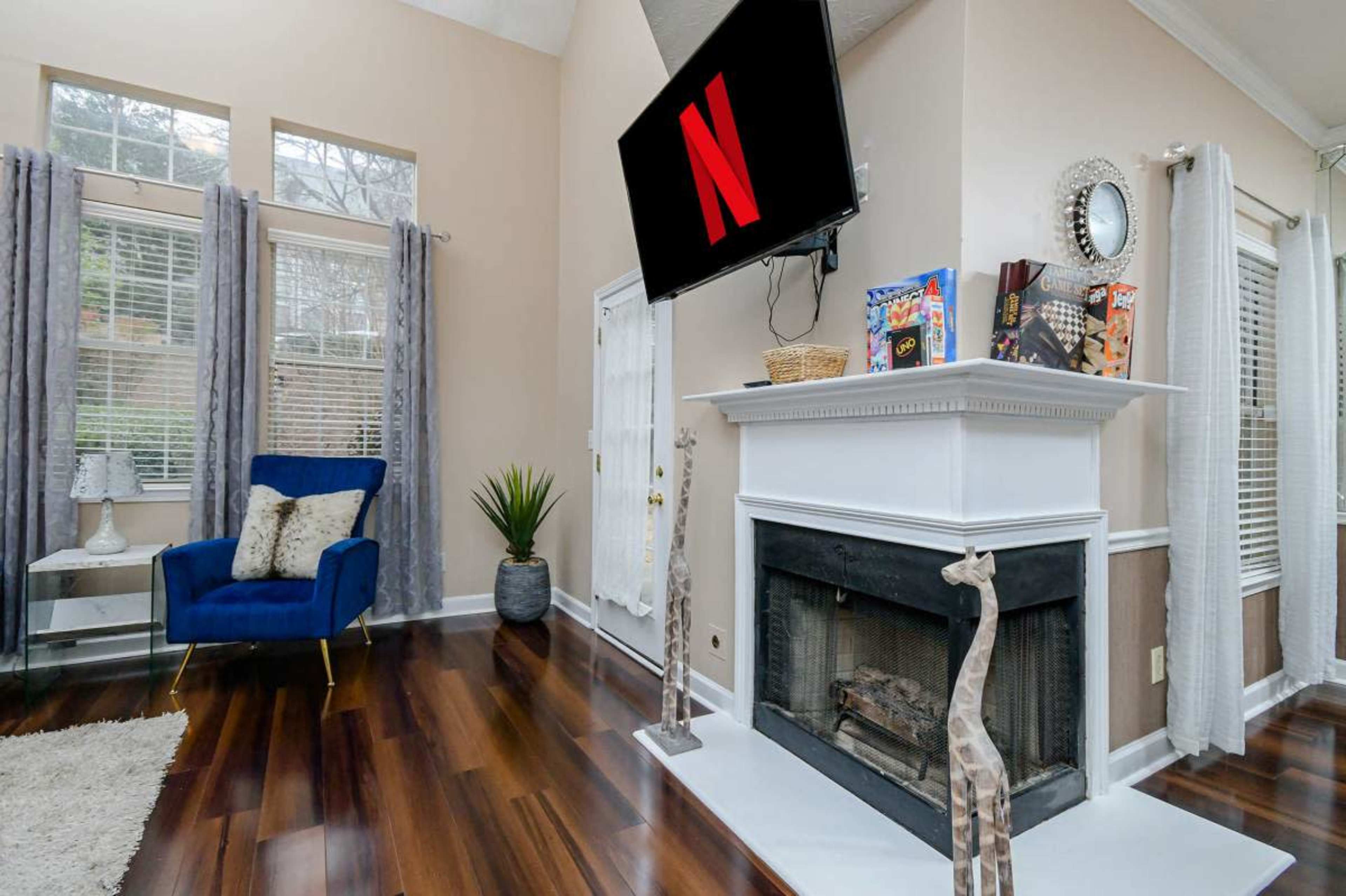 A living room features a blue armchair, a television mounted above a white fireplace, and decorative giraffe sculptures on either side of the mantel.