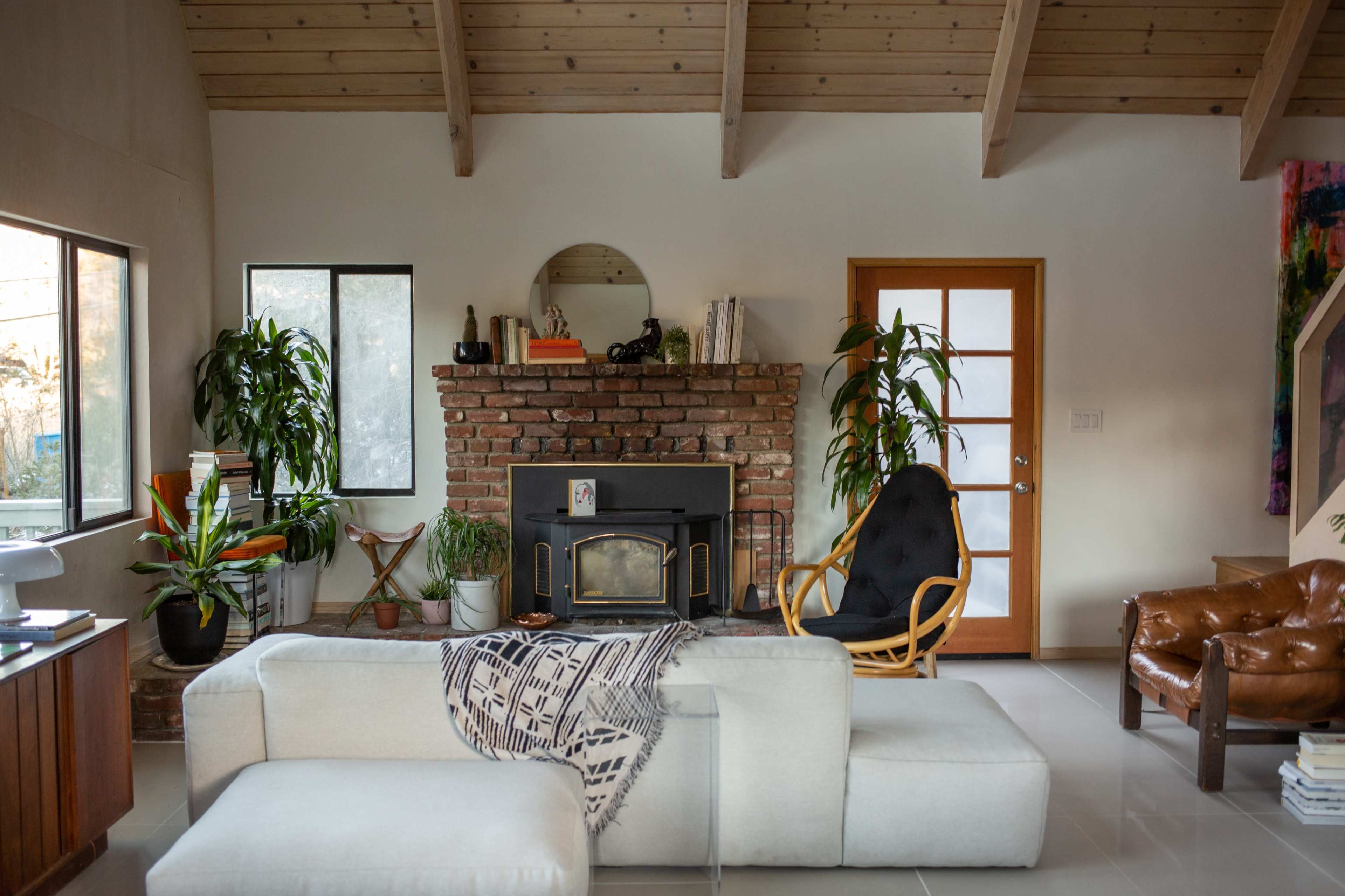 A modern living room featuring a brick fireplace, large windows, a beige sectional sofa, and various indoor plants.