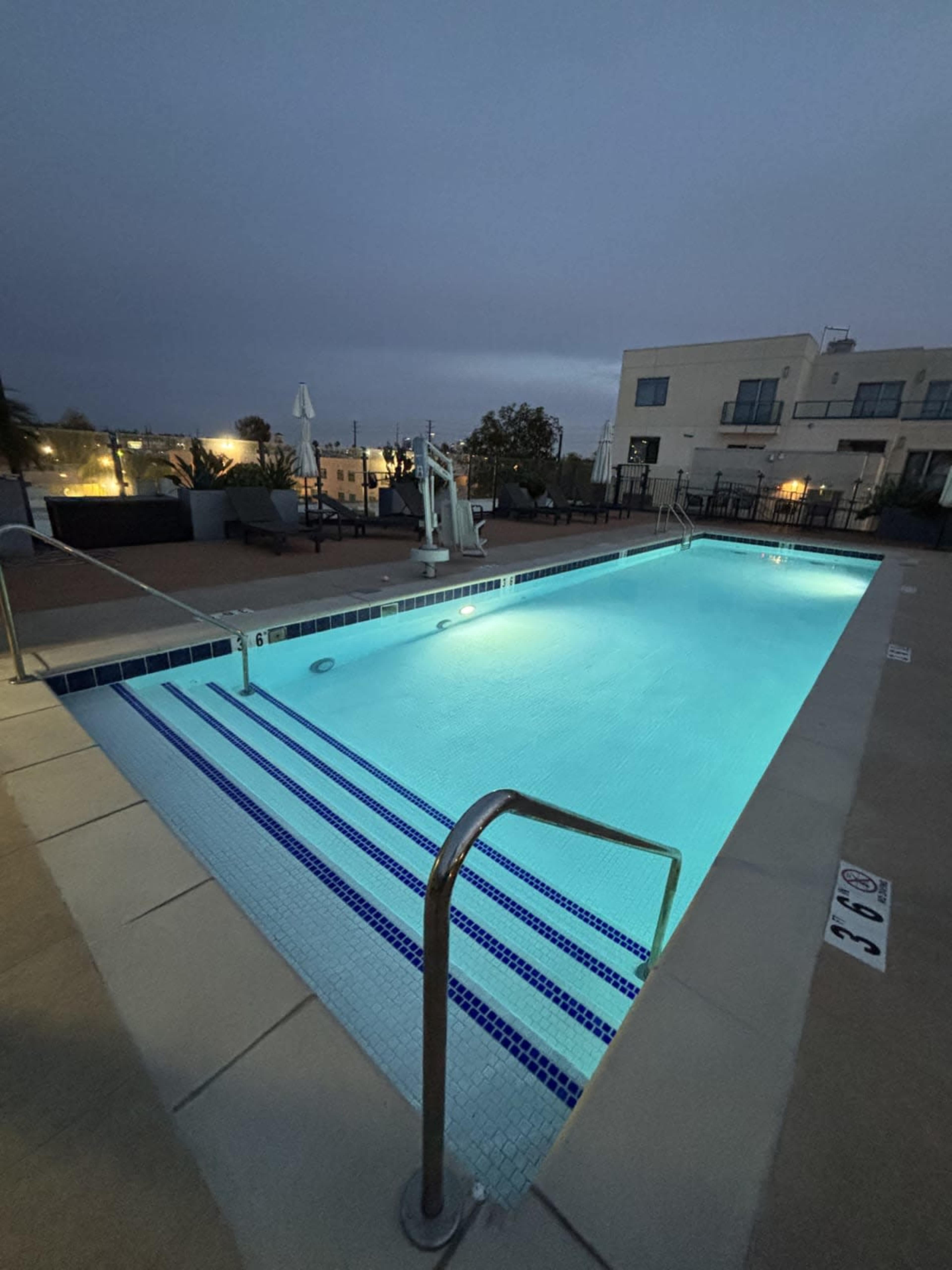 A well-lit swimming pool with a ladder is set against a dusky sky and surrounded by a paved area and lounge chairs.