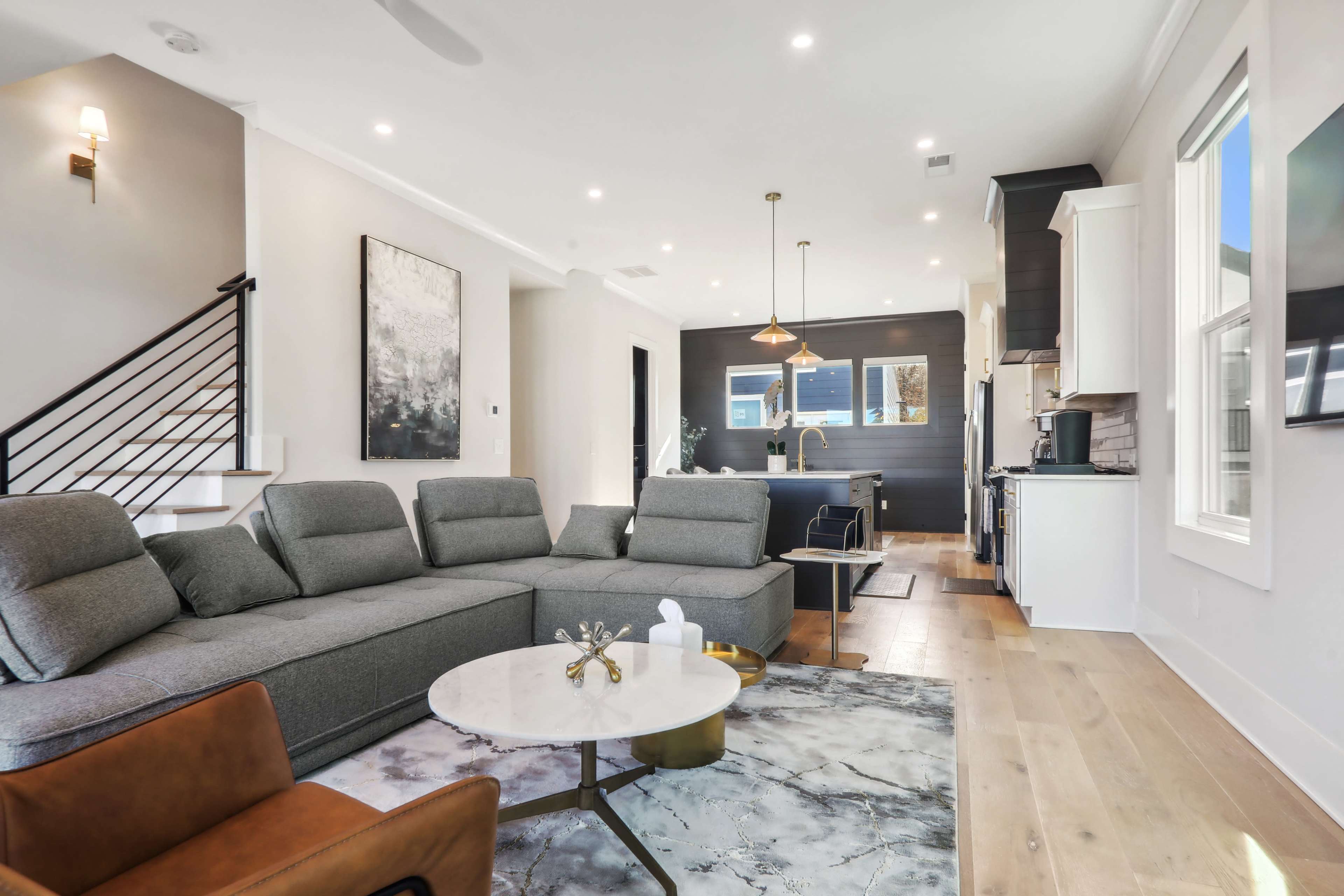 A modern living room features a gray sectional sofa, a round coffee table, and a view of a kitchen in the background.