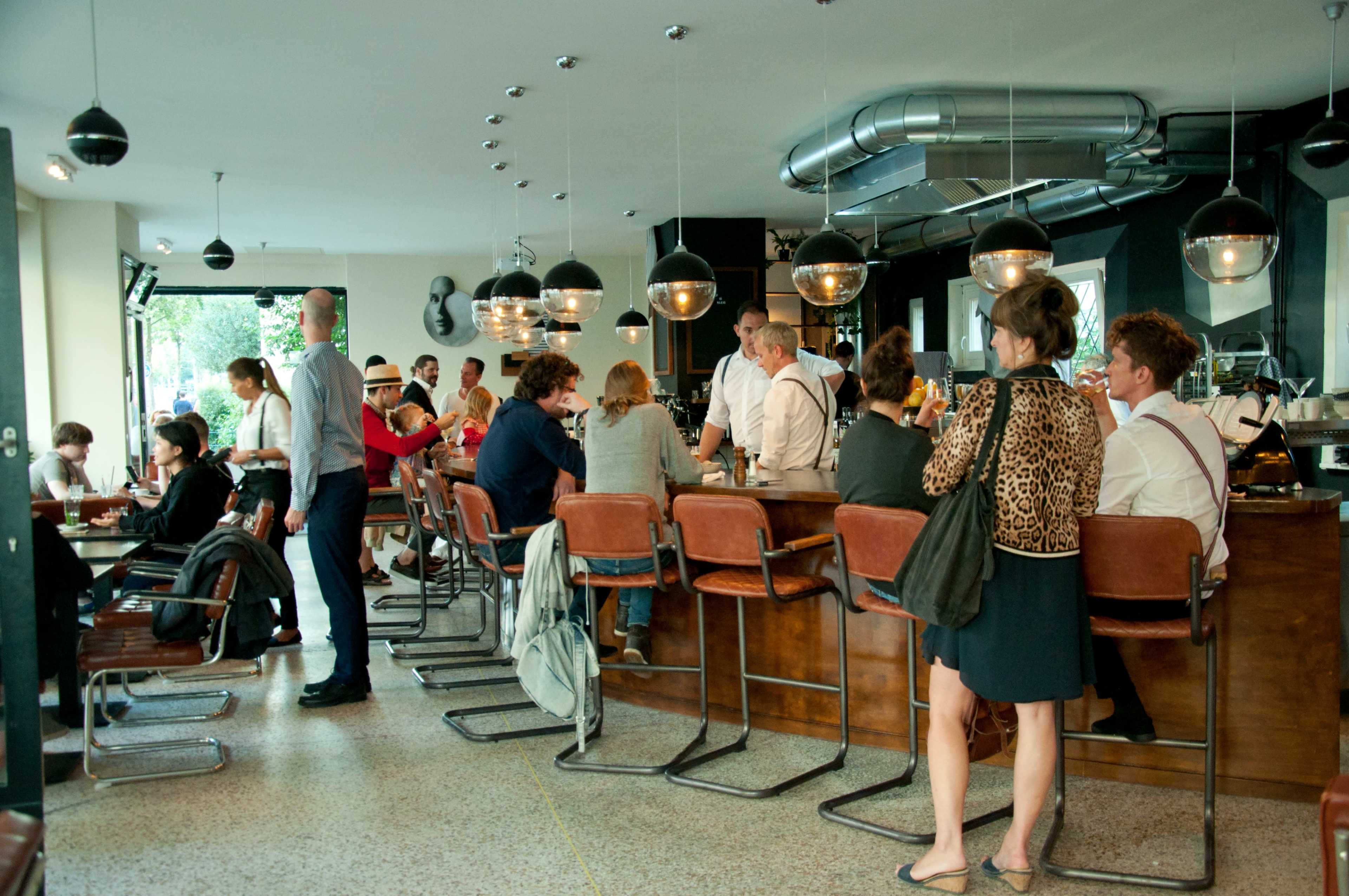 A bustling cafe features patrons seated at a bar and tables, with a mix of people engaged in conversation and enjoying drinks.