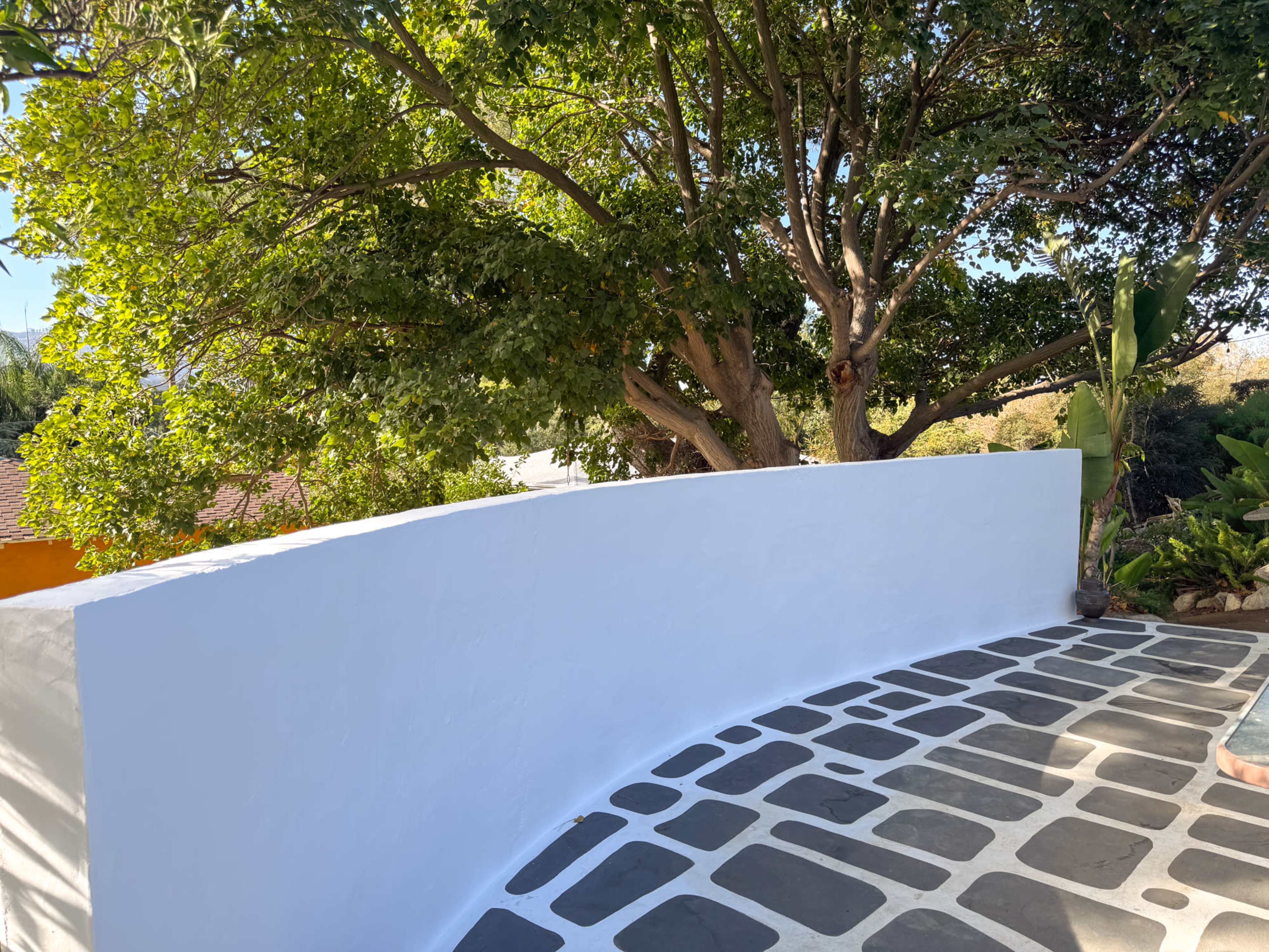 A curved white wall with a stone floor is set against a backdrop of lush green trees.