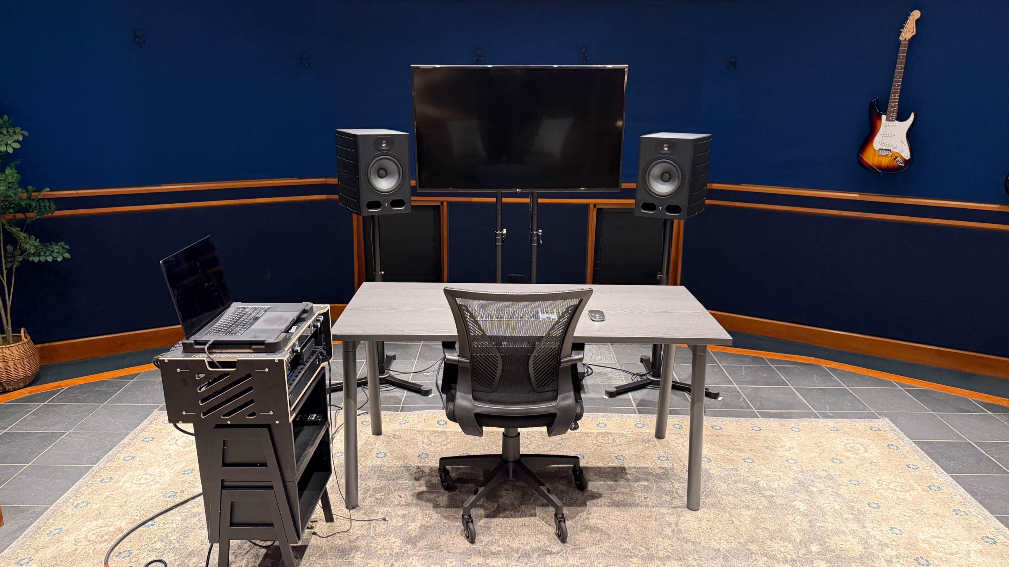 A modern audio studio features a desk, a chair, two speakers, a television on a stand, and a laptop on a wheeled rack, all set against a dark blue backdrop.