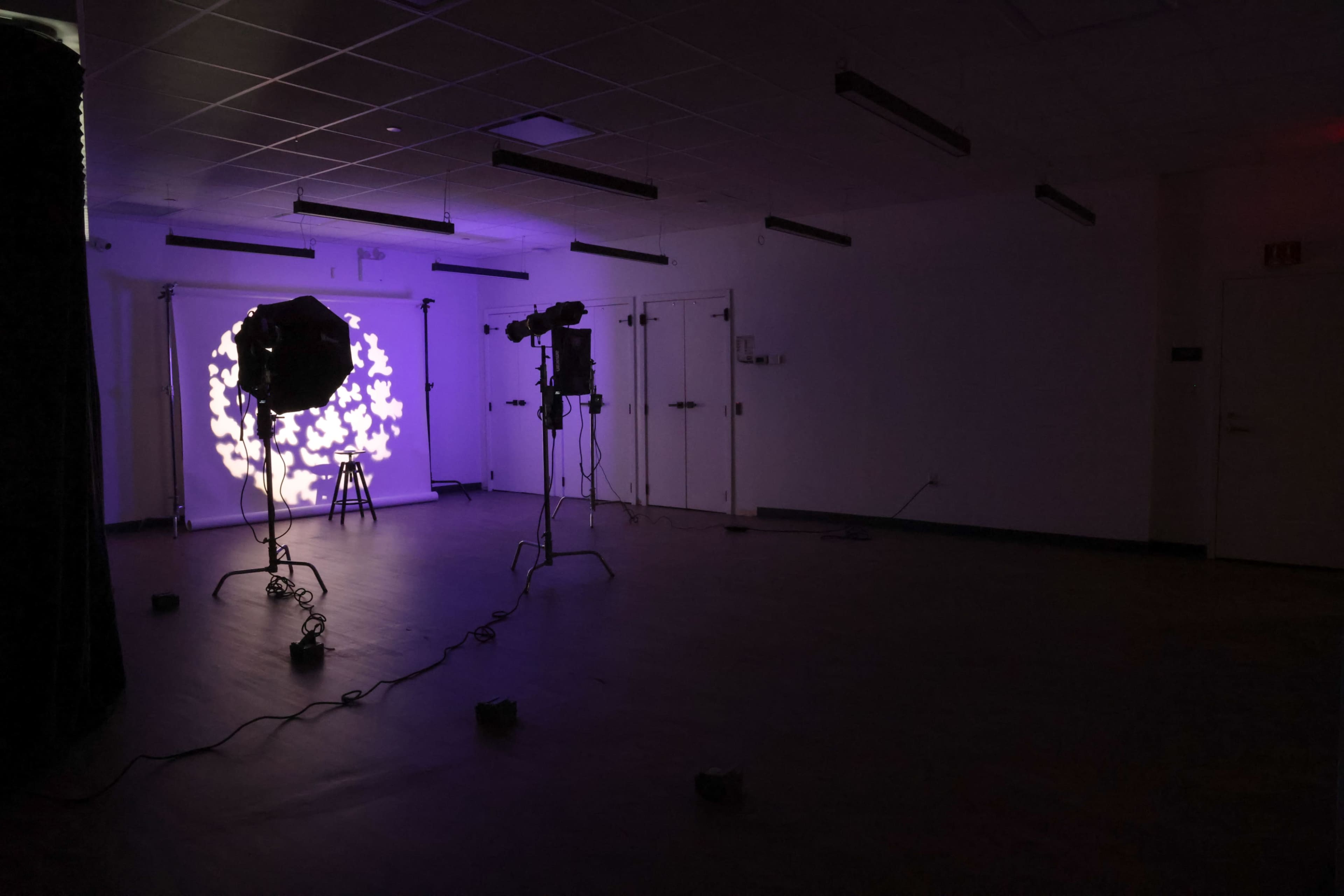 A dimly lit studio space features two lighting setups pointing at a backdrop, with colored light patterns projected on the wall.