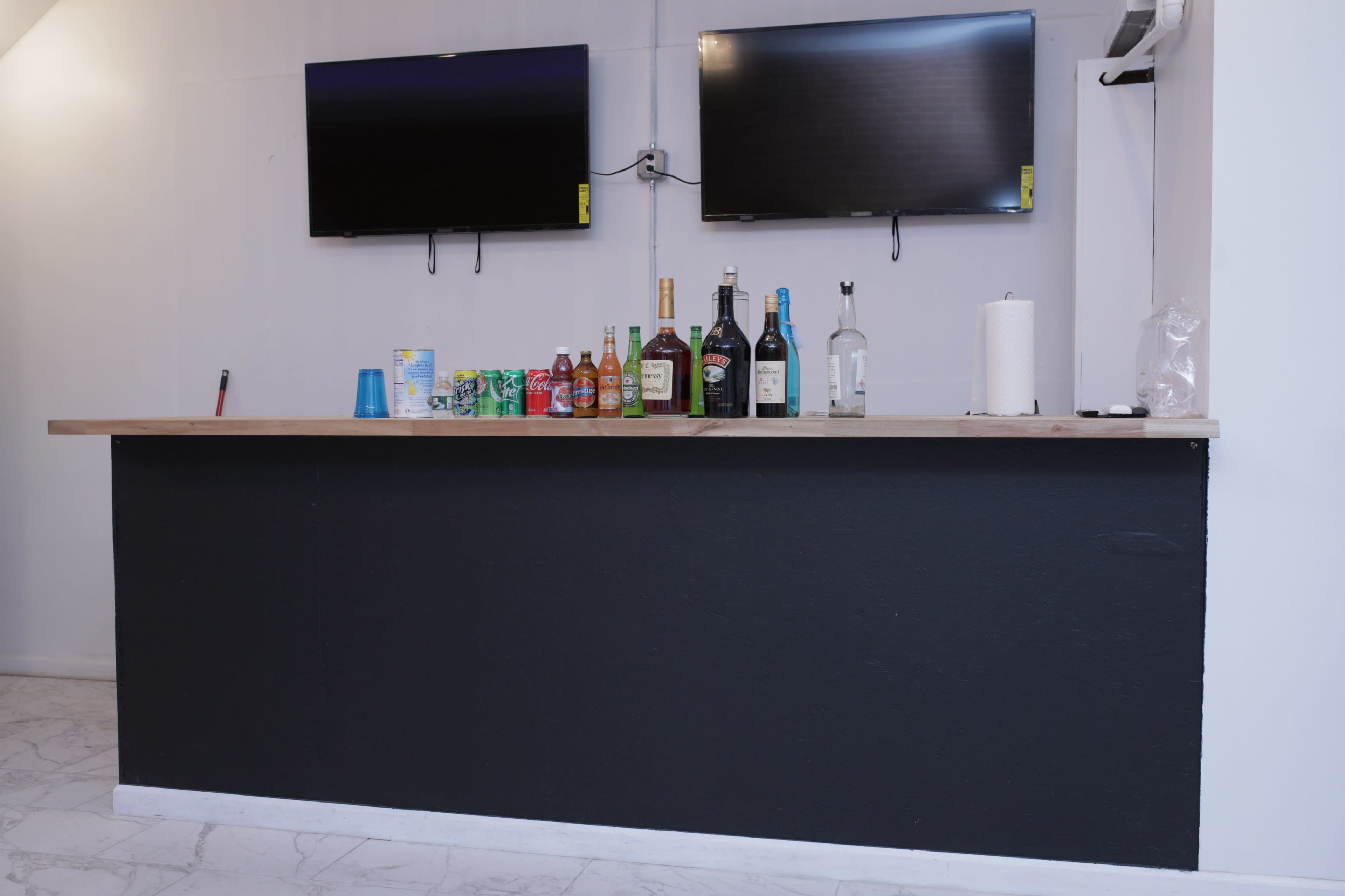 A bar counter with two mounted TVs and various bottles of beverages is set against a white wall.