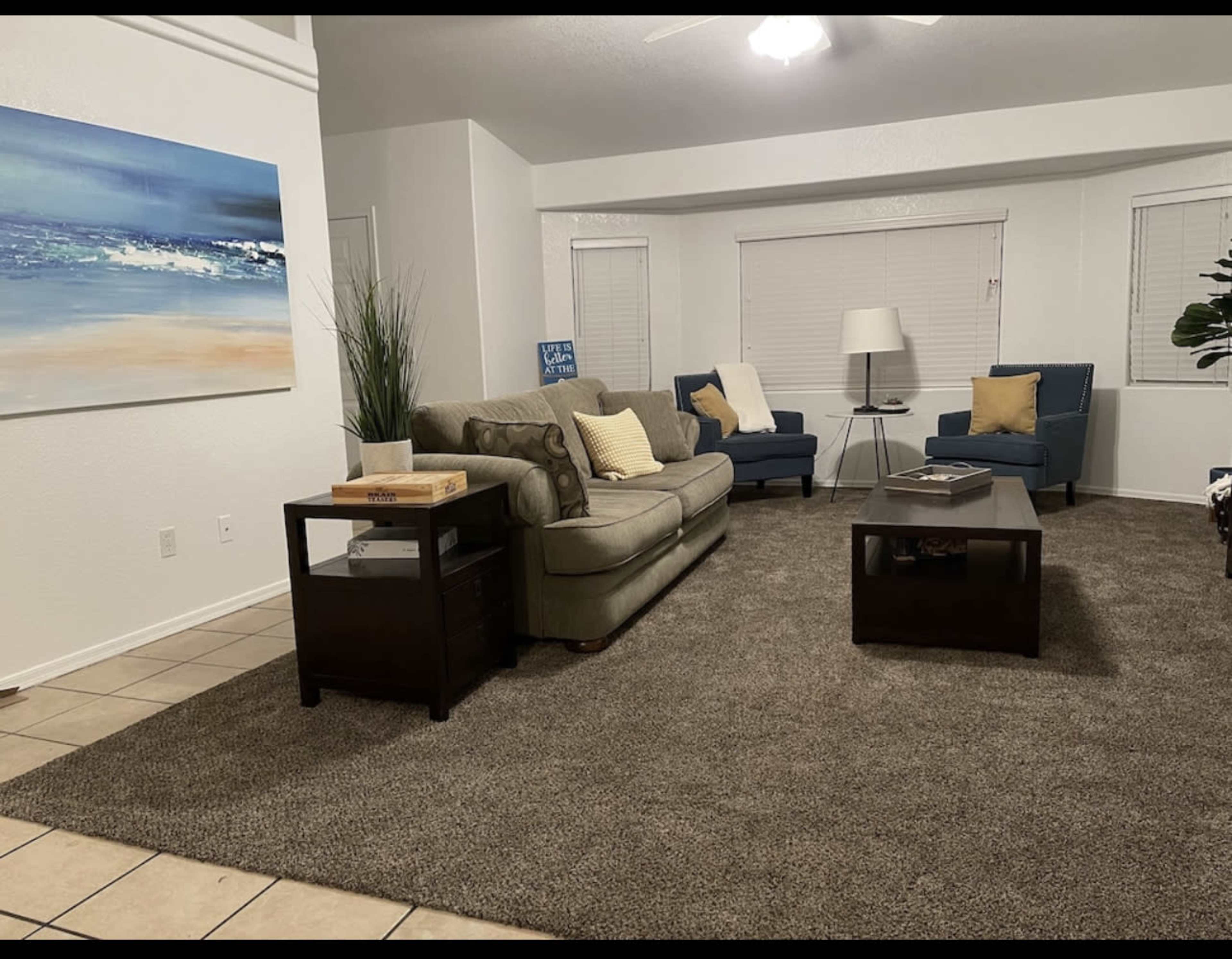 The image shows a living room with a beige sofa, two blue armchairs, a dark wooden coffee table, and a large painting of a seascape on the wall.