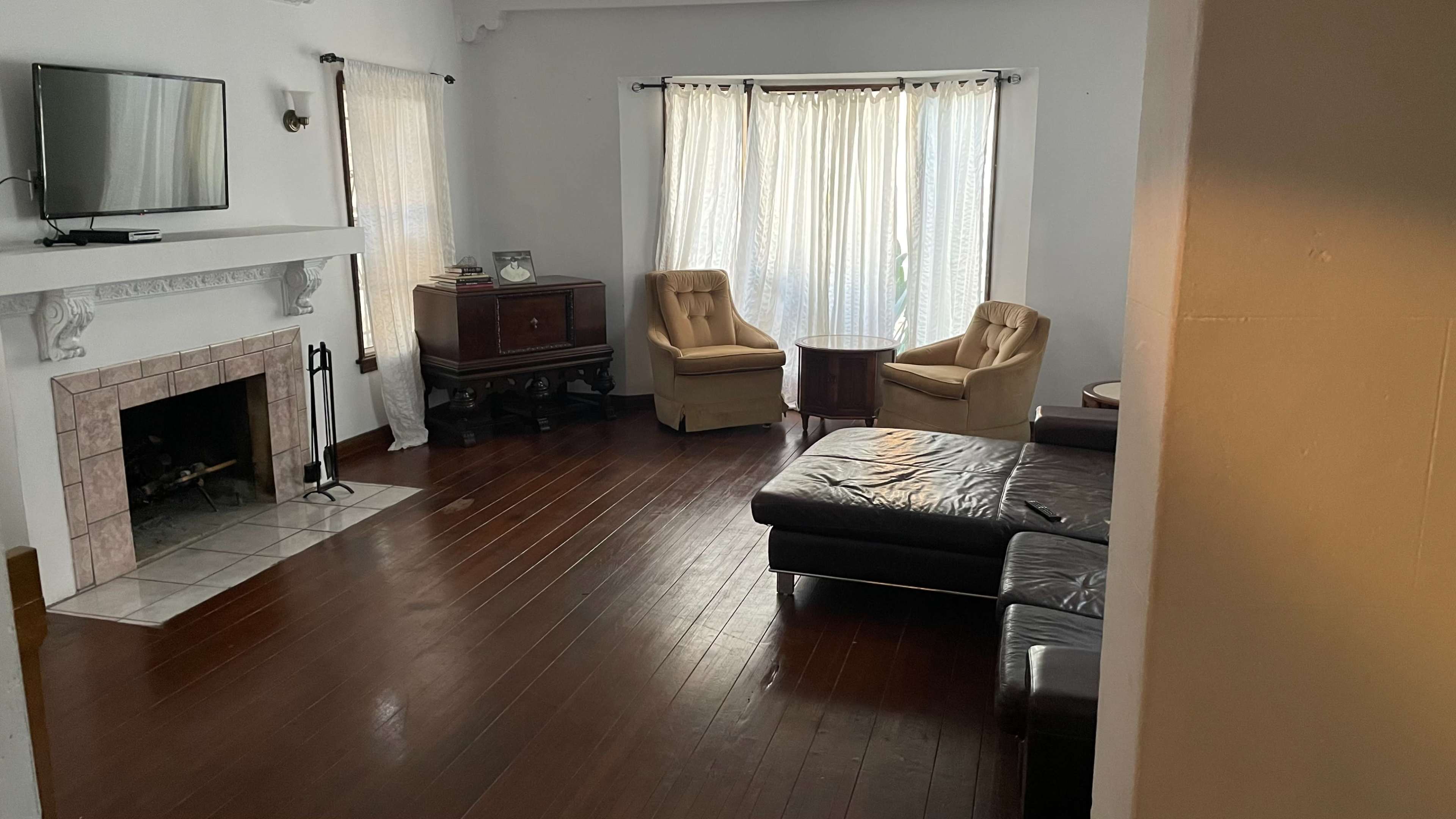A living room features a television on the wall, two armchairs, a small table, and a leather sectional sofa, with natural light streaming through sheer curtains.