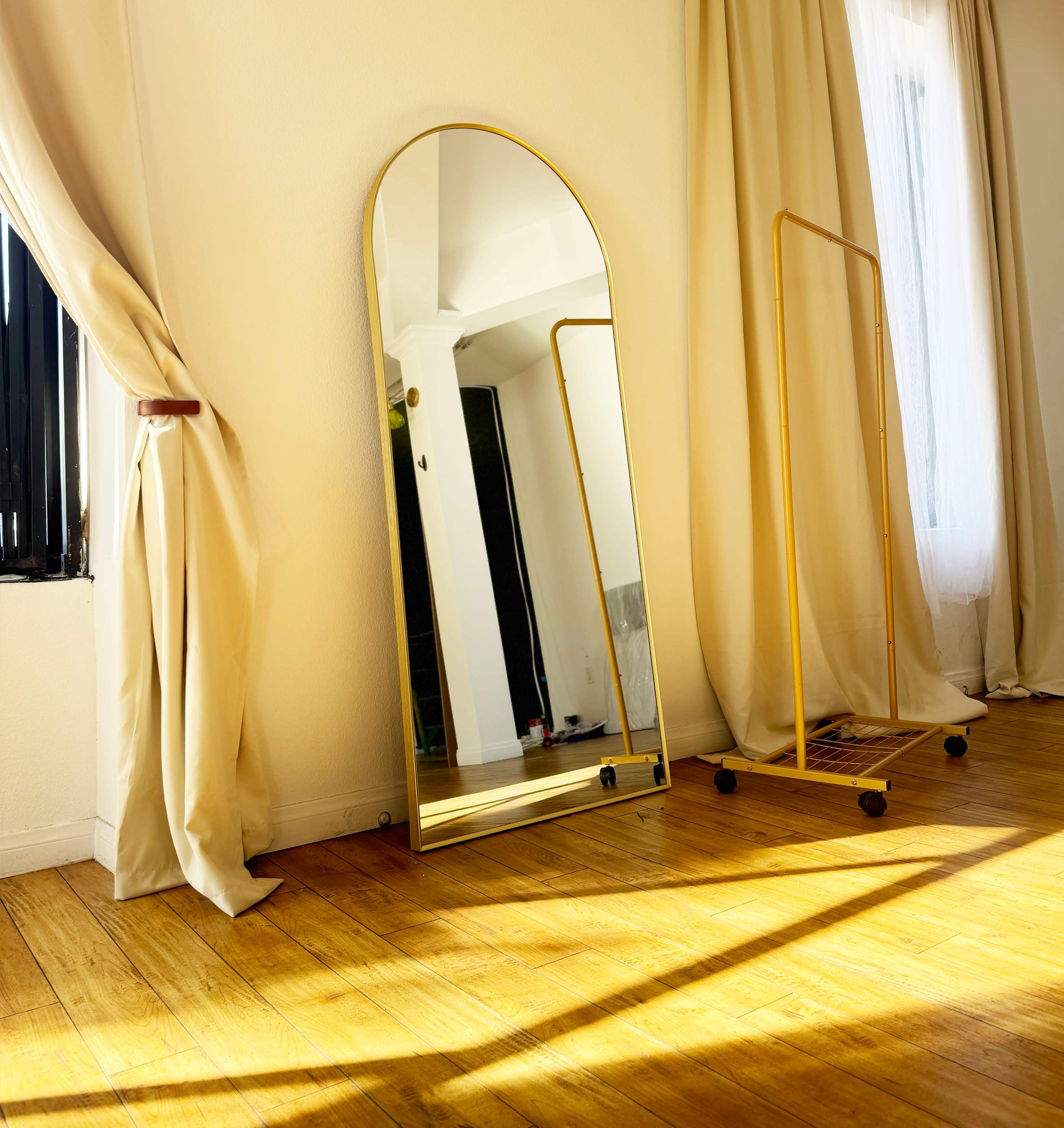 A large arched mirror stands against a wall in a sunlit room, with a golden frame and wooden floorboards beneath it.