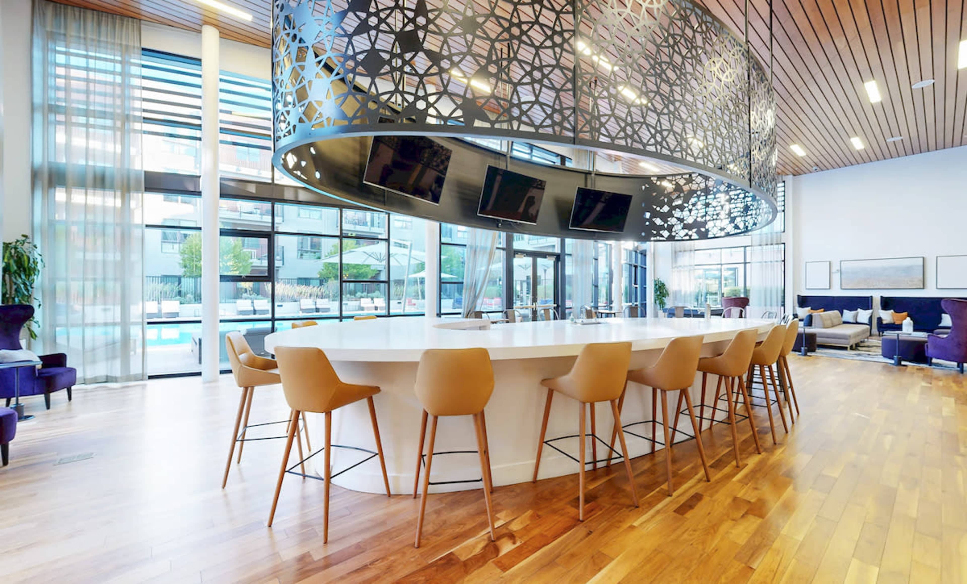 The image shows a modern lounge area with a circular bar, surrounded by high stools, large windows letting in natural light, and a view of an outdoor pool.