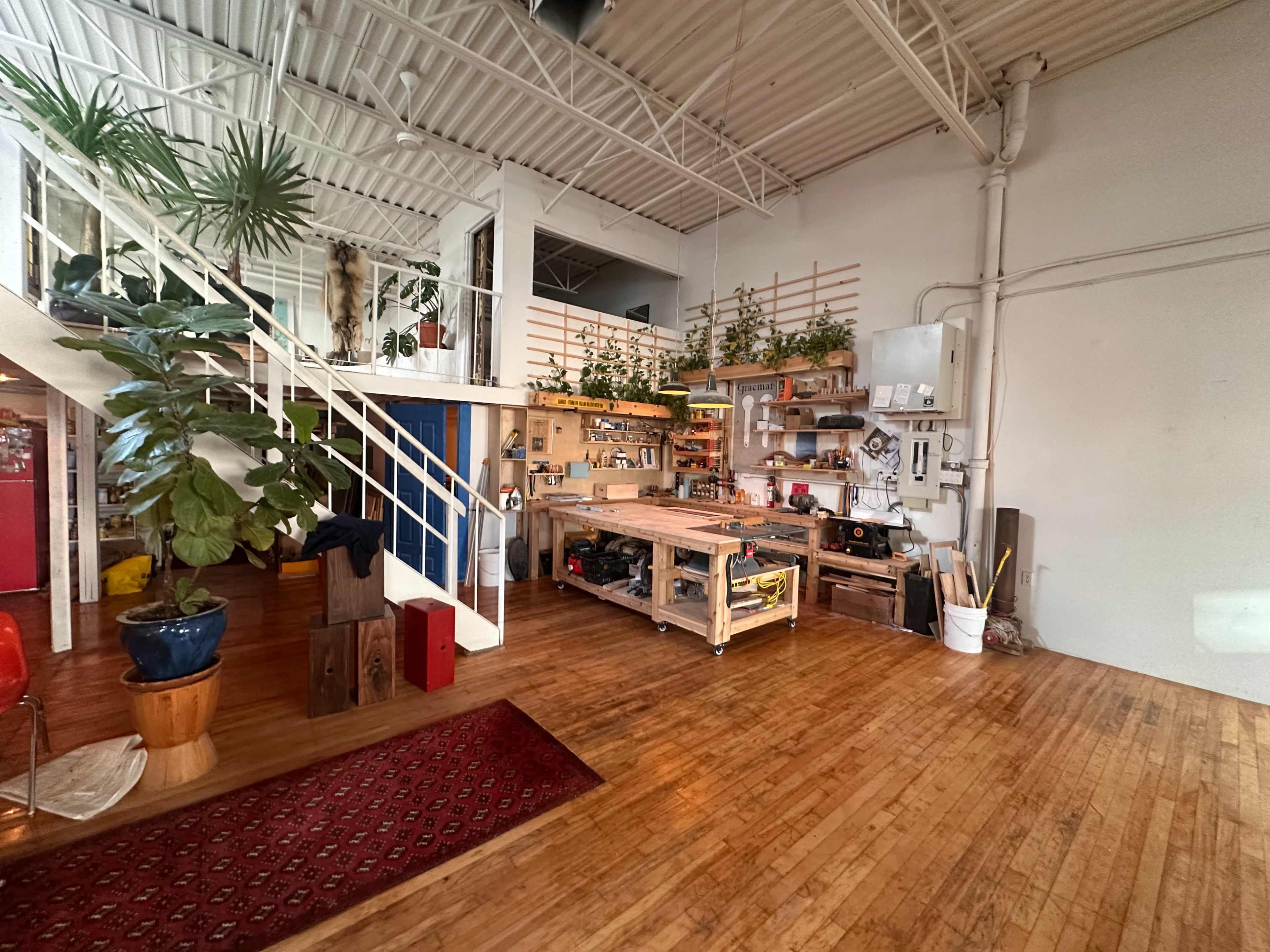 The image depicts a spacious interior with hardwood flooring, featuring a workshop area with various tools, plants, and a staircase leading to an upper level.