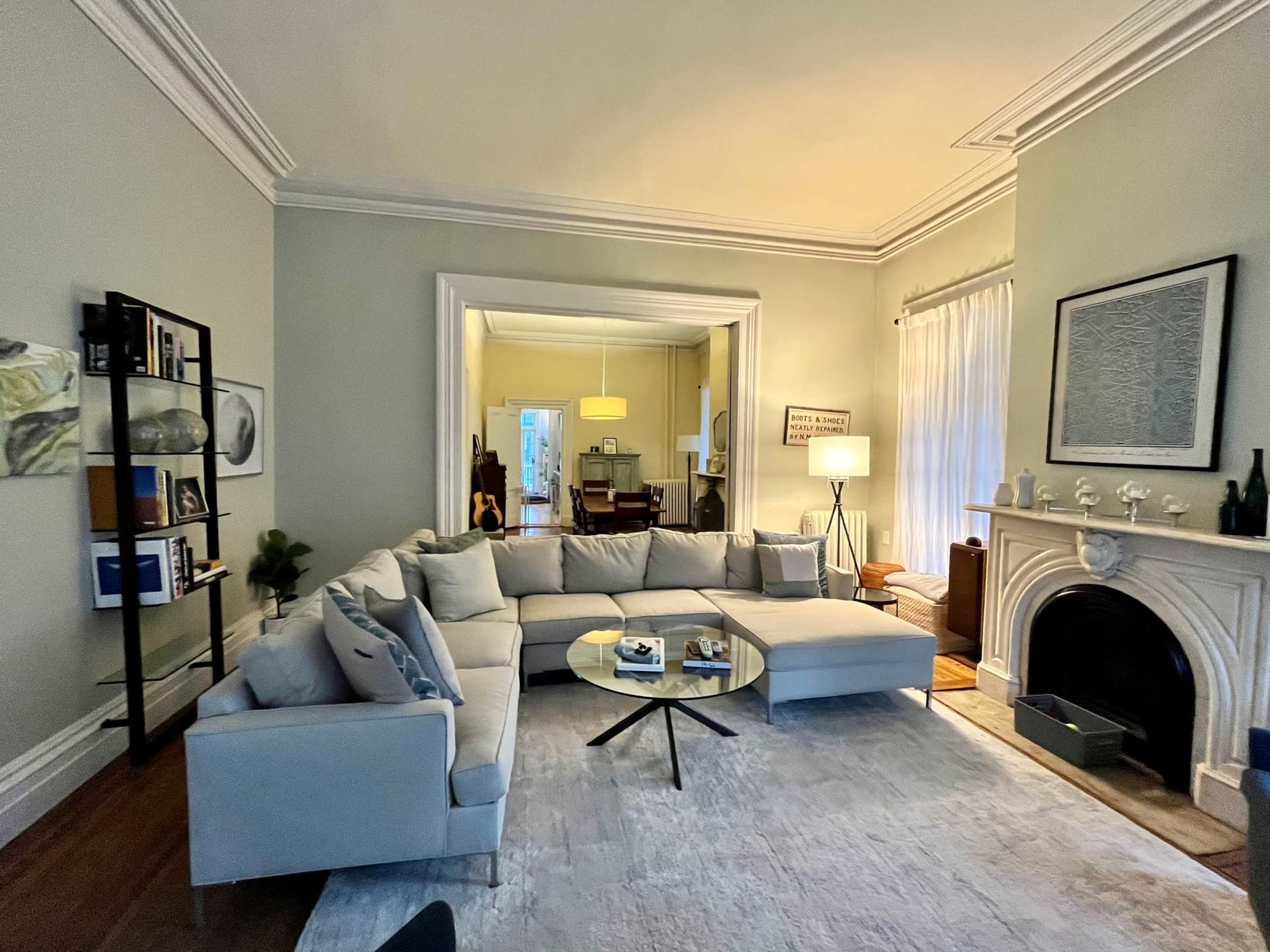 A light-colored living room features a large sectional sofa, a round coffee table, a black bookshelf, and a fireplace, with natural light streaming in from nearby windows.