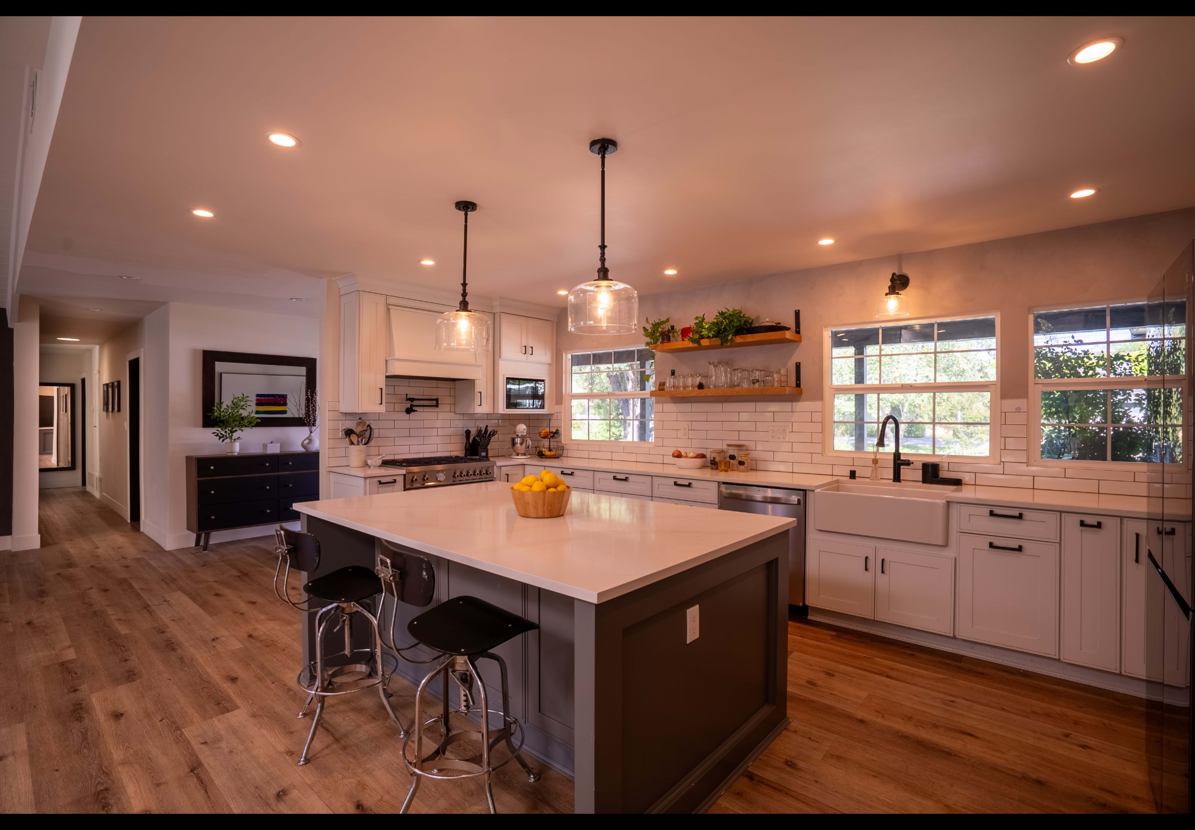The image shows a modern kitchen featuring a central island with bar stools, white cabinets, and large windows that allow natural light to fill the space.
