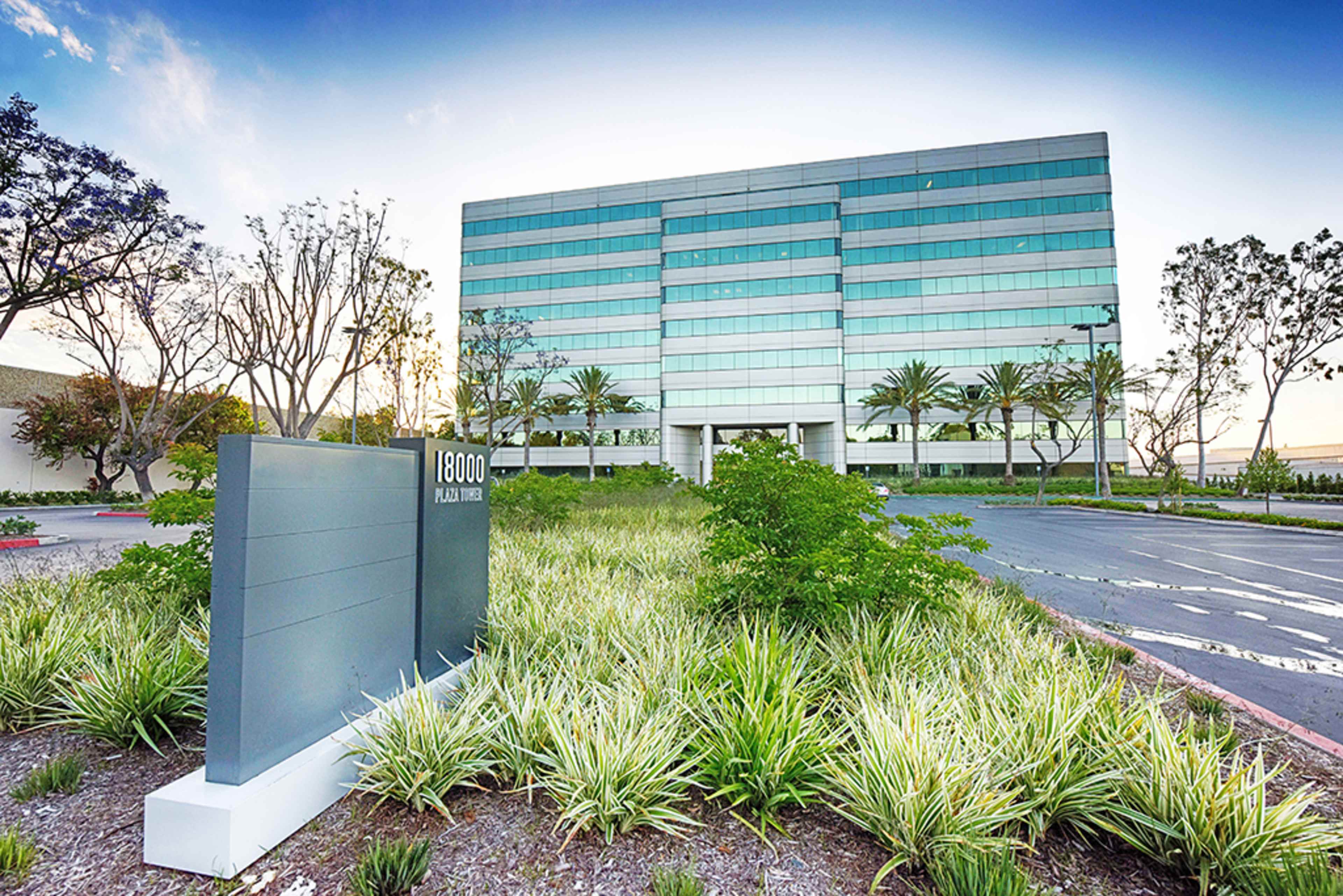A modern six-story glass office building surrounded by landscaped greenery and a sign displaying "18000 Plaza Tower."