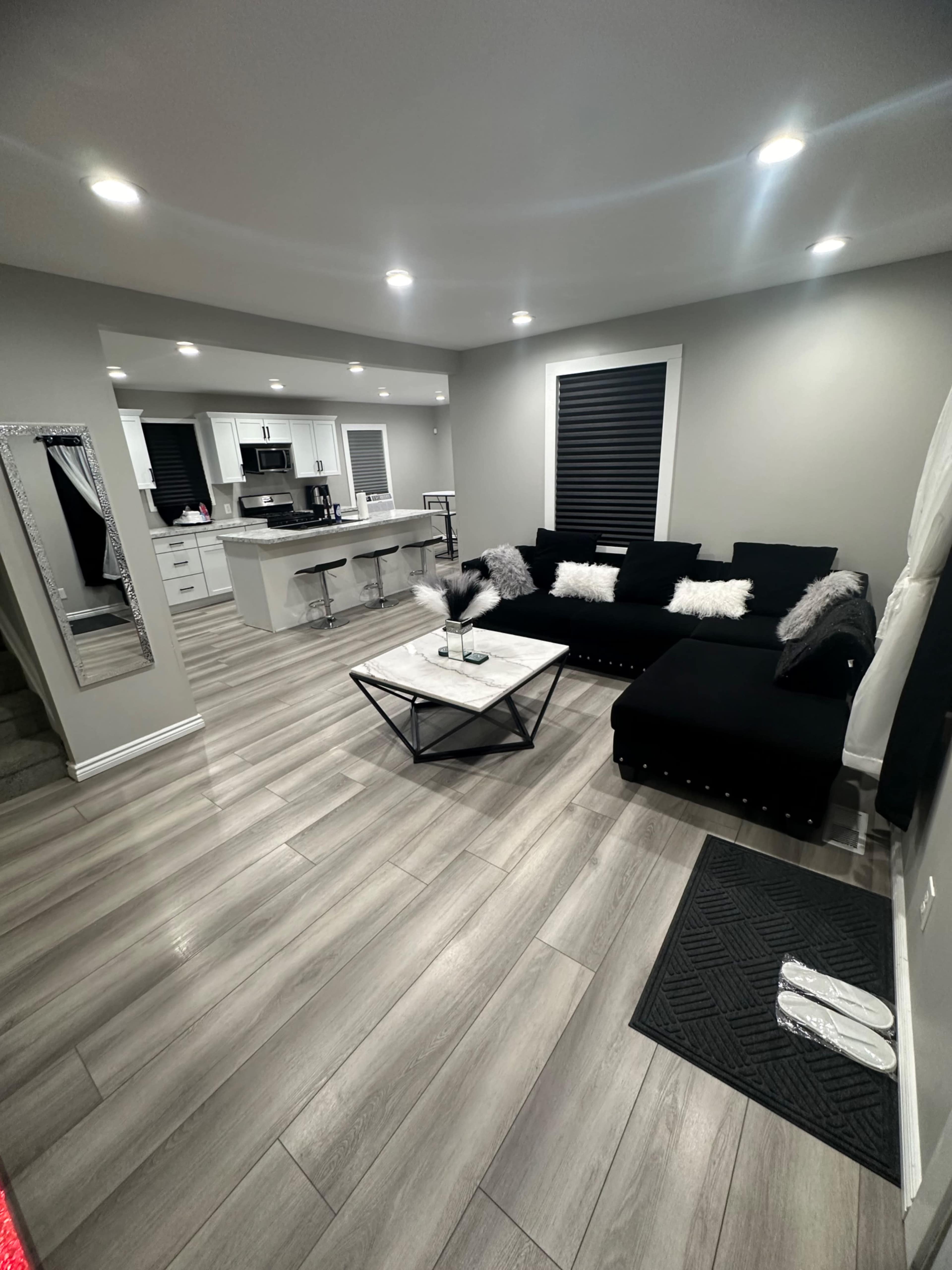The image shows a modern living room with a black sectional sofa, a marble coffee table, and a kitchen area visible in the background.