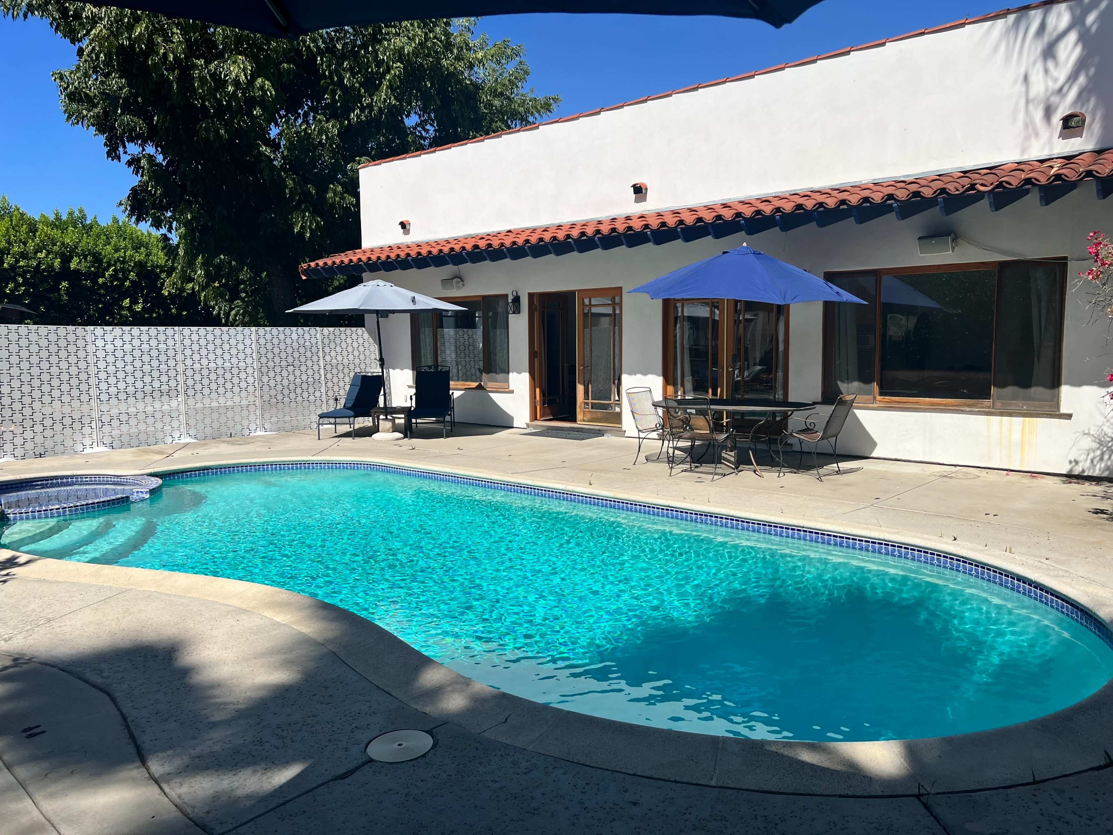 The image shows a backyard with a swimming pool, lounge chairs, and a dining table shaded by umbrellas, set against a sunny blue sky.