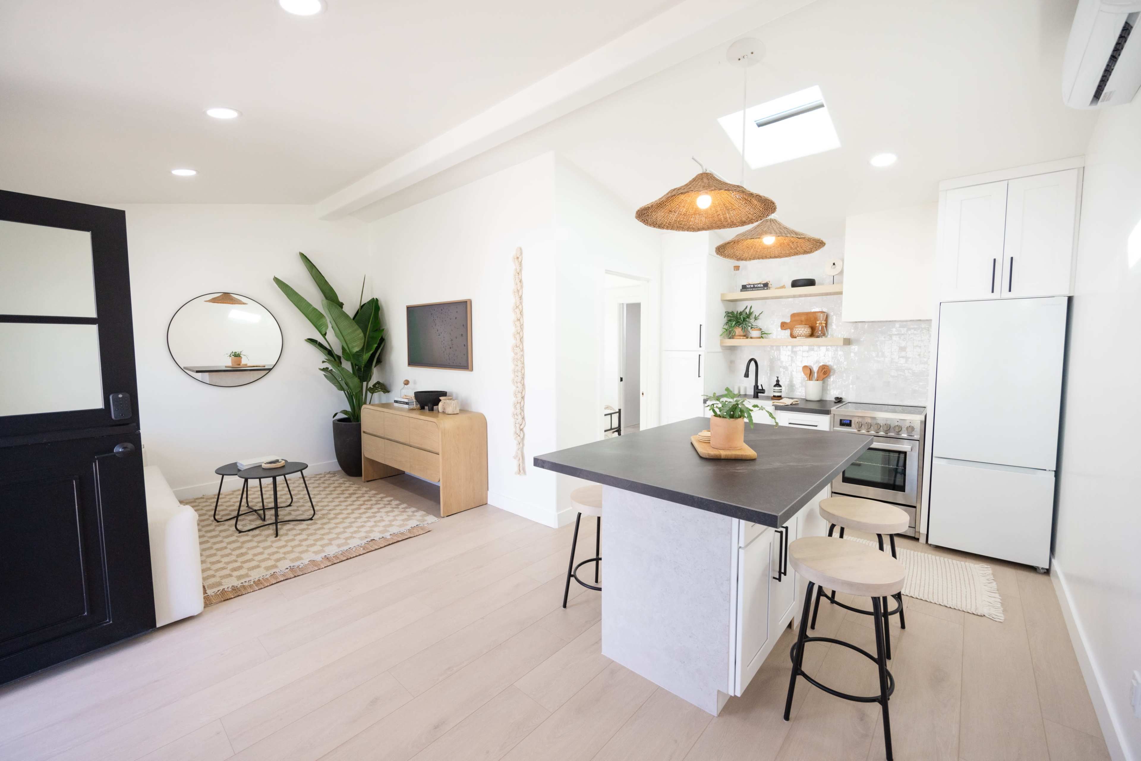 A modern kitchen and living area featuring a central island with bar stools, a light wood sideboard, plants, and large windows.
