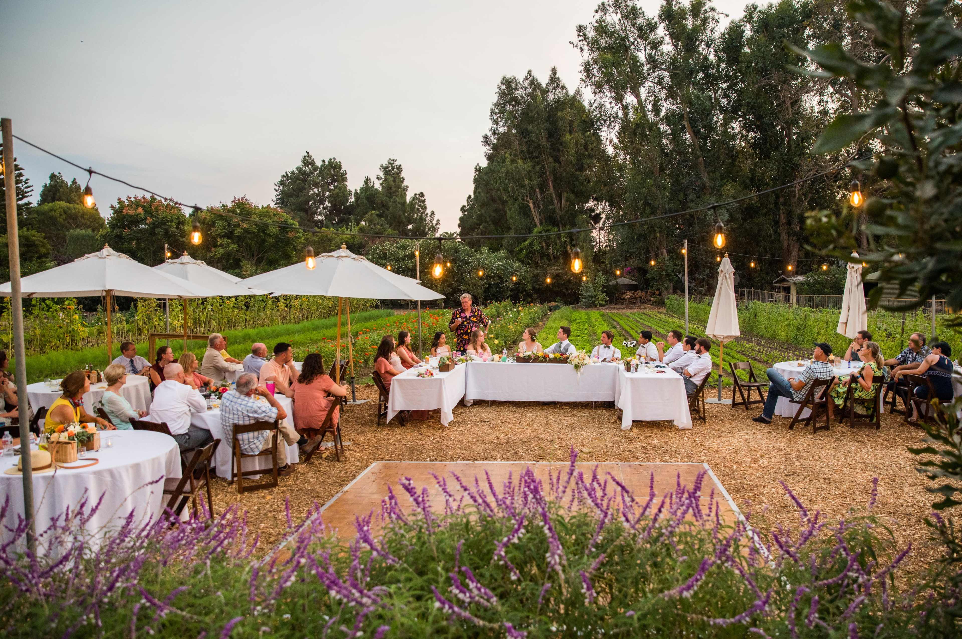 A group of people sit at several long tables under string lights in an outdoor setting, surrounded by greenery and flowering plants.