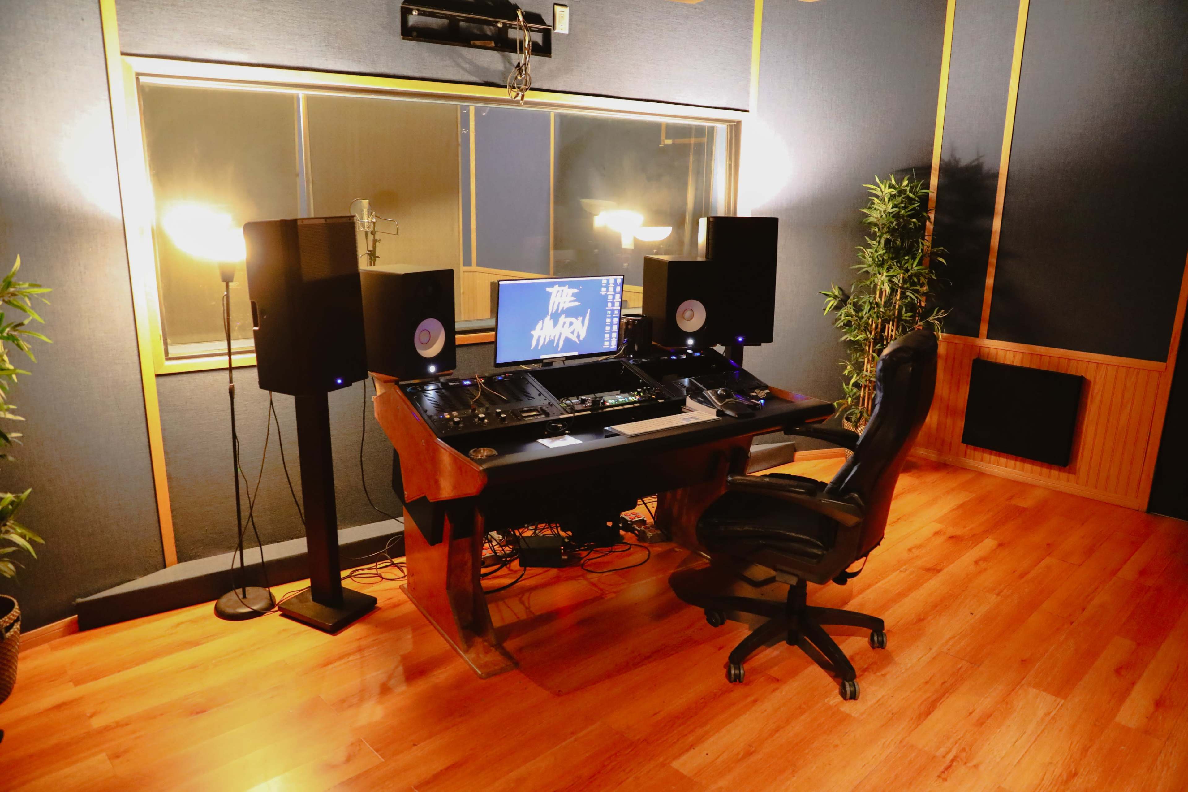 The image shows a recording studio with a central desk equipped with audio equipment, a computer monitor, and studio speakers, surrounded by acoustic panels and plants.