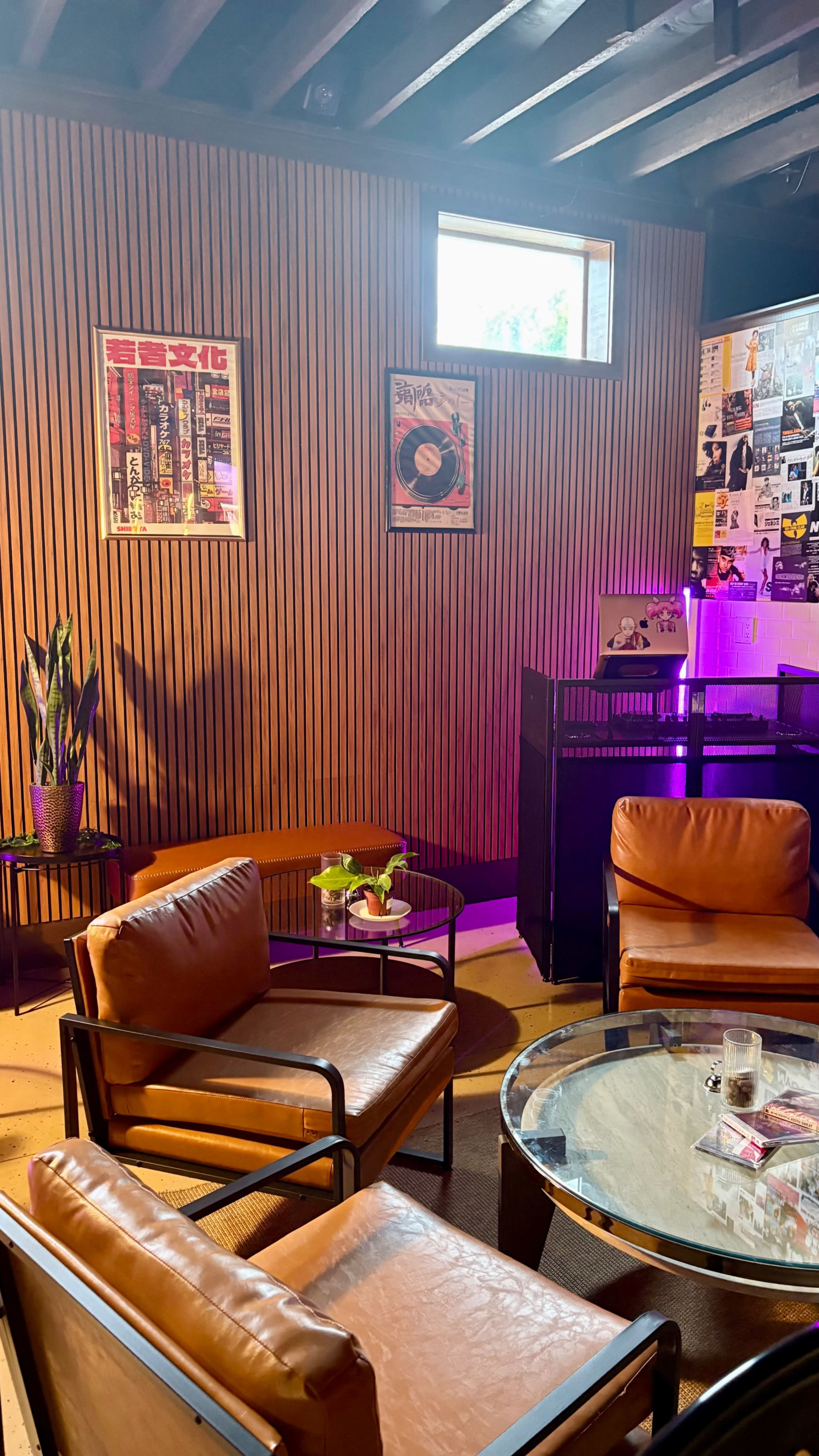 The image shows a cozy indoor space featuring leather armchairs, a glass coffee table, wooden paneling on the walls, and colorful posters.