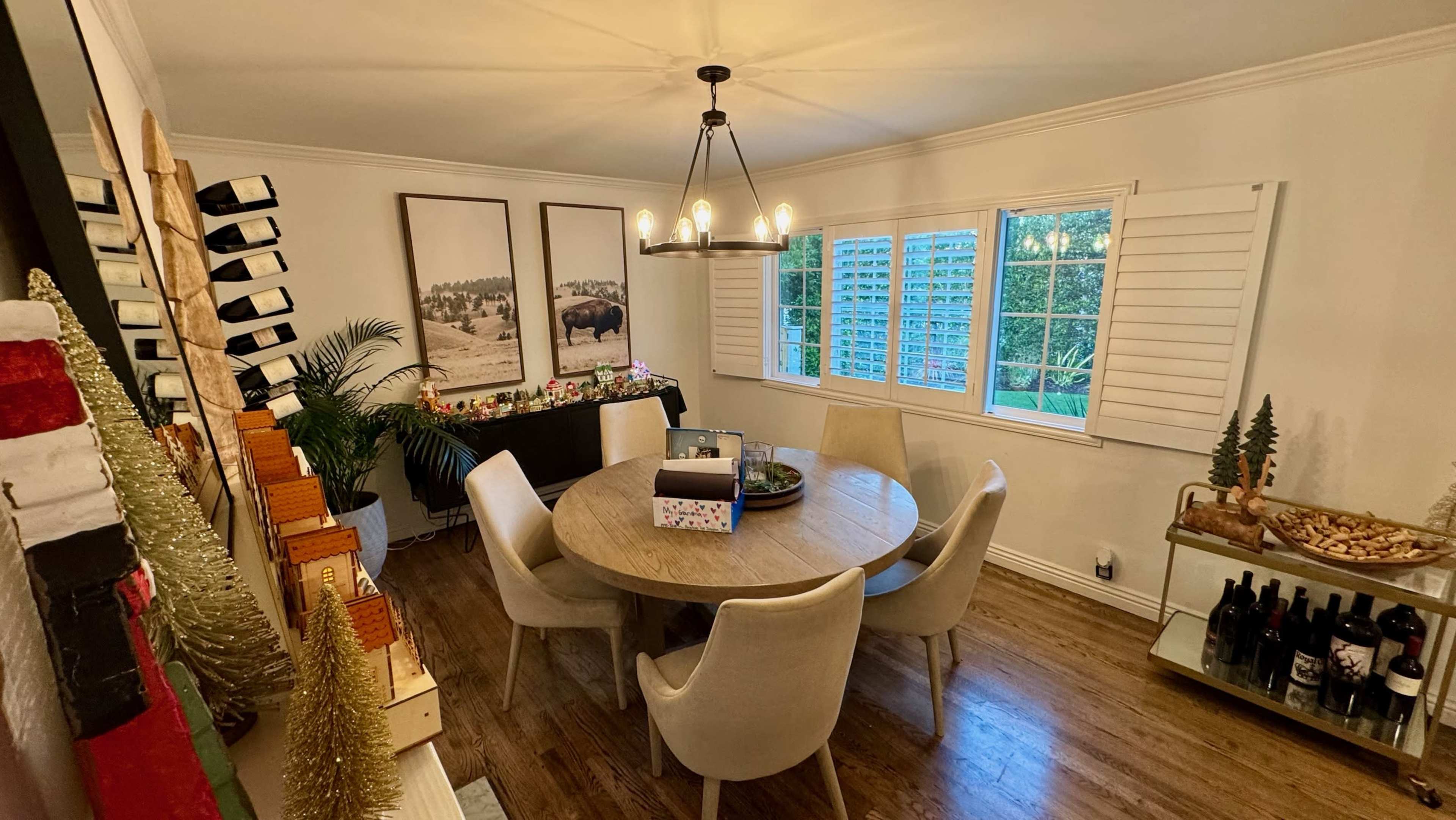 A round dining table with six chairs is set in a well-lit room featuring large windows, decorative artwork, and a side table with plants and wine.