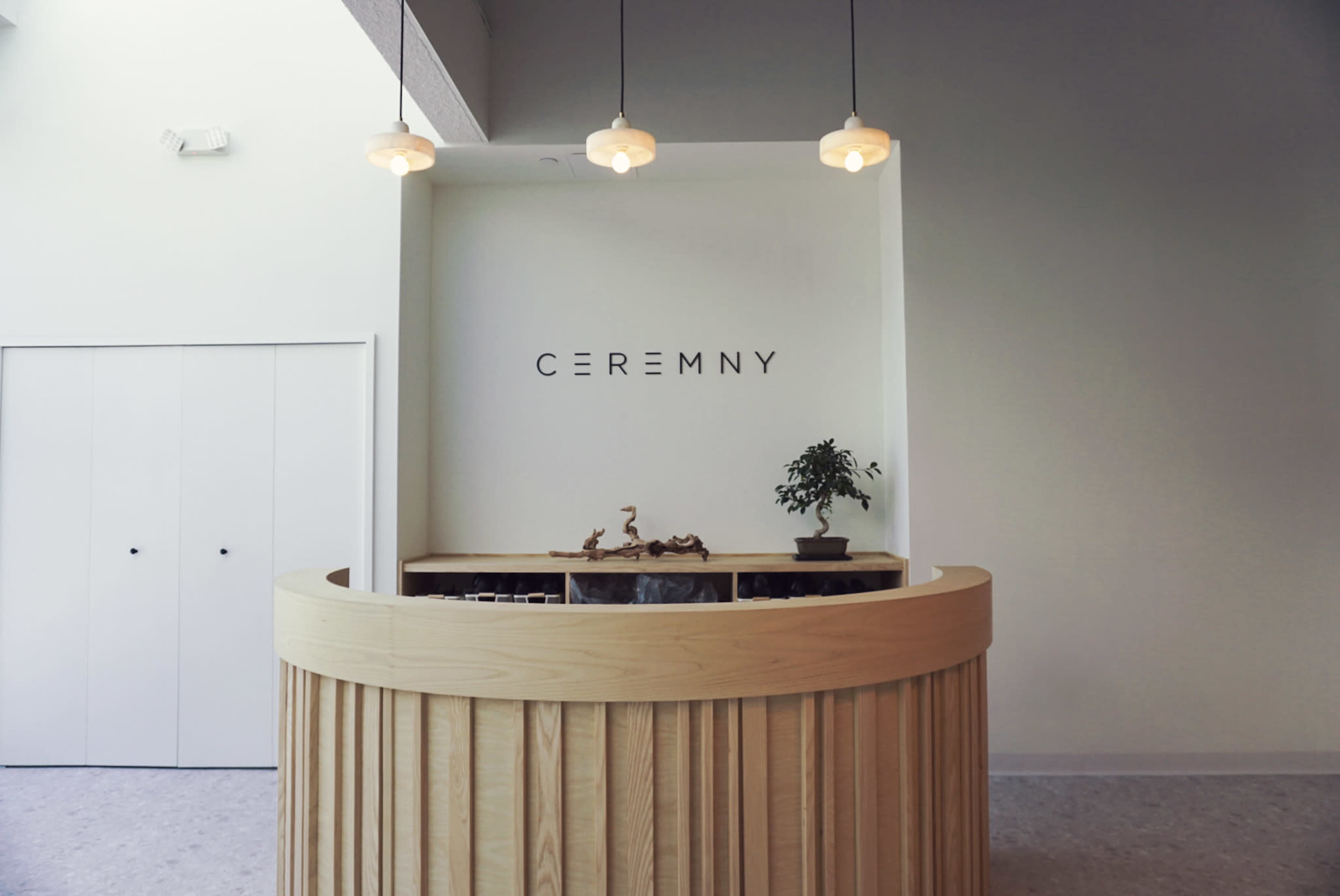 A curved wooden reception desk with the word "CEREMONY" displayed on the wall behind it is illuminated by pendant lights in a minimalist interior.