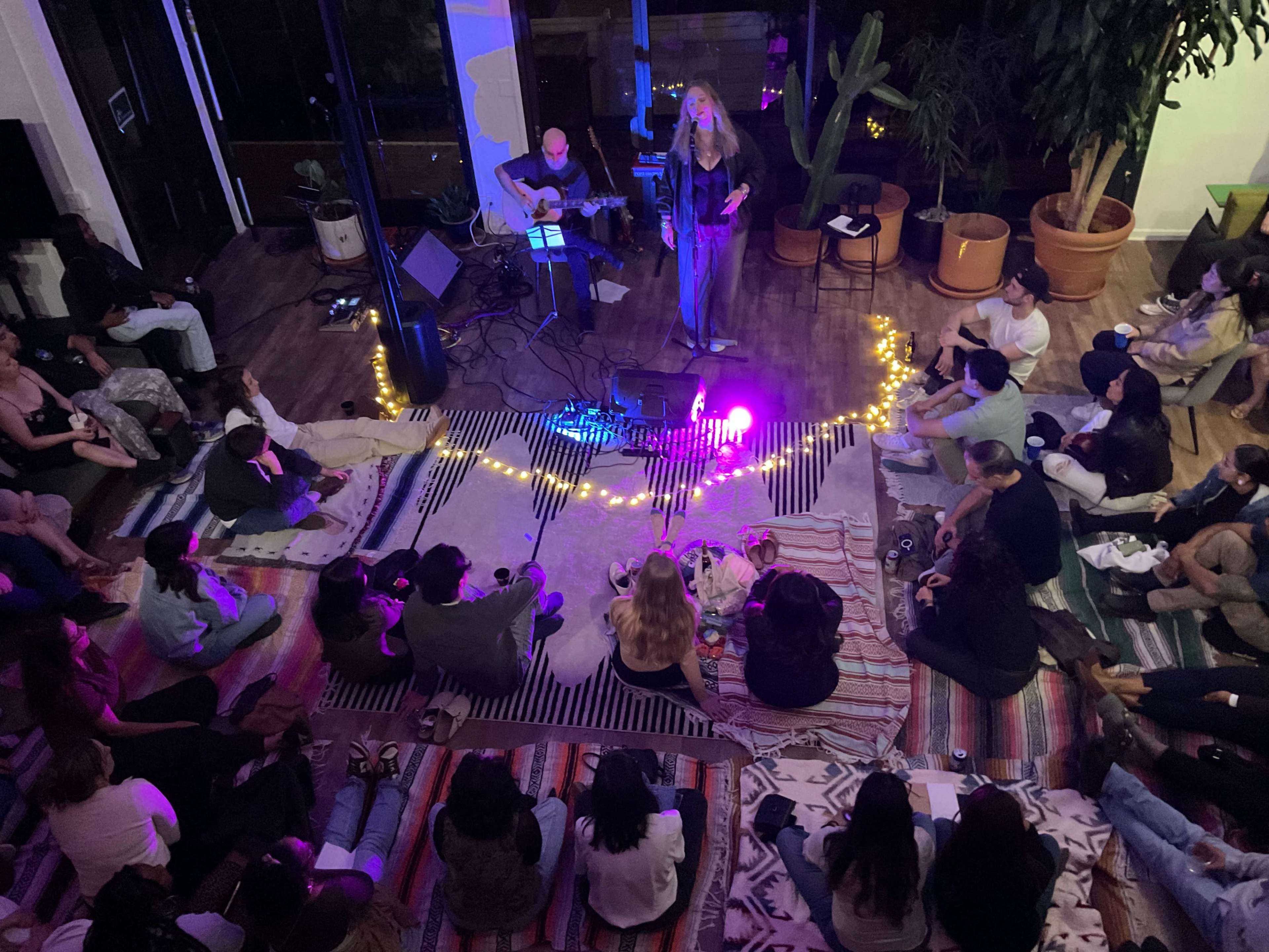 A singer performs on stage with a guitarist while an audience sits on the floor, surrounded by decorative lights and plants.