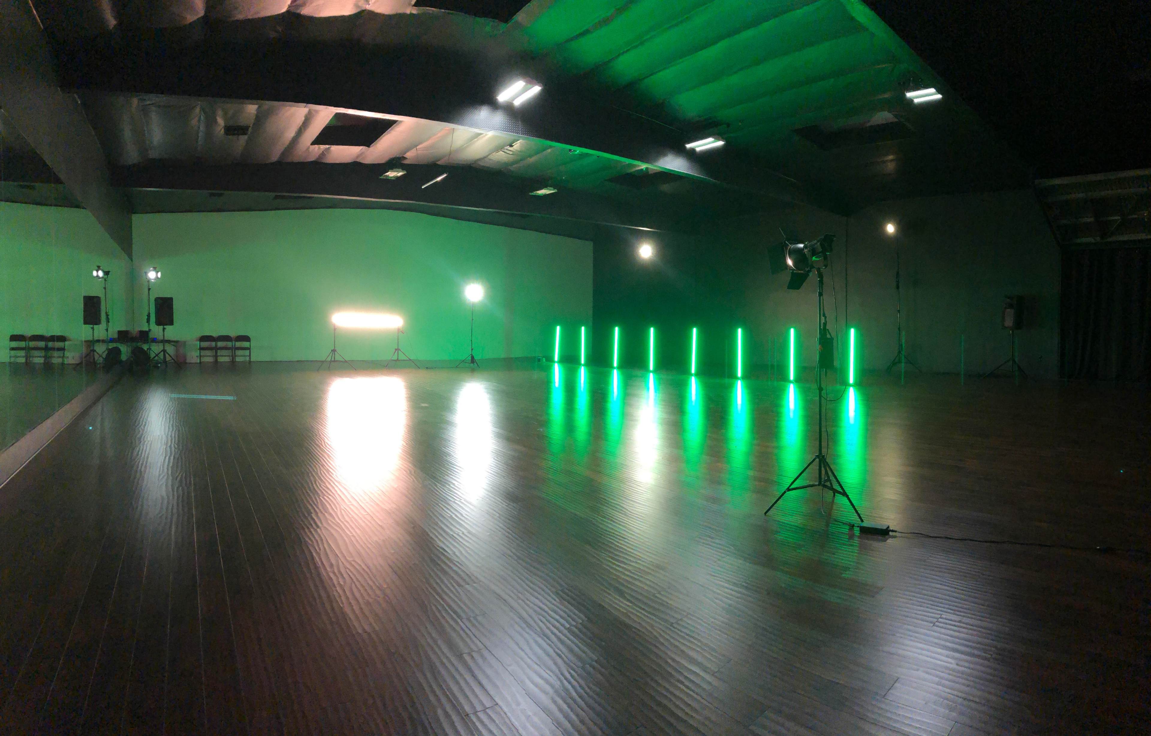 The image shows a spacious indoor studio with green lighting effects, featuring a row of illuminated vertical poles and several light stands.