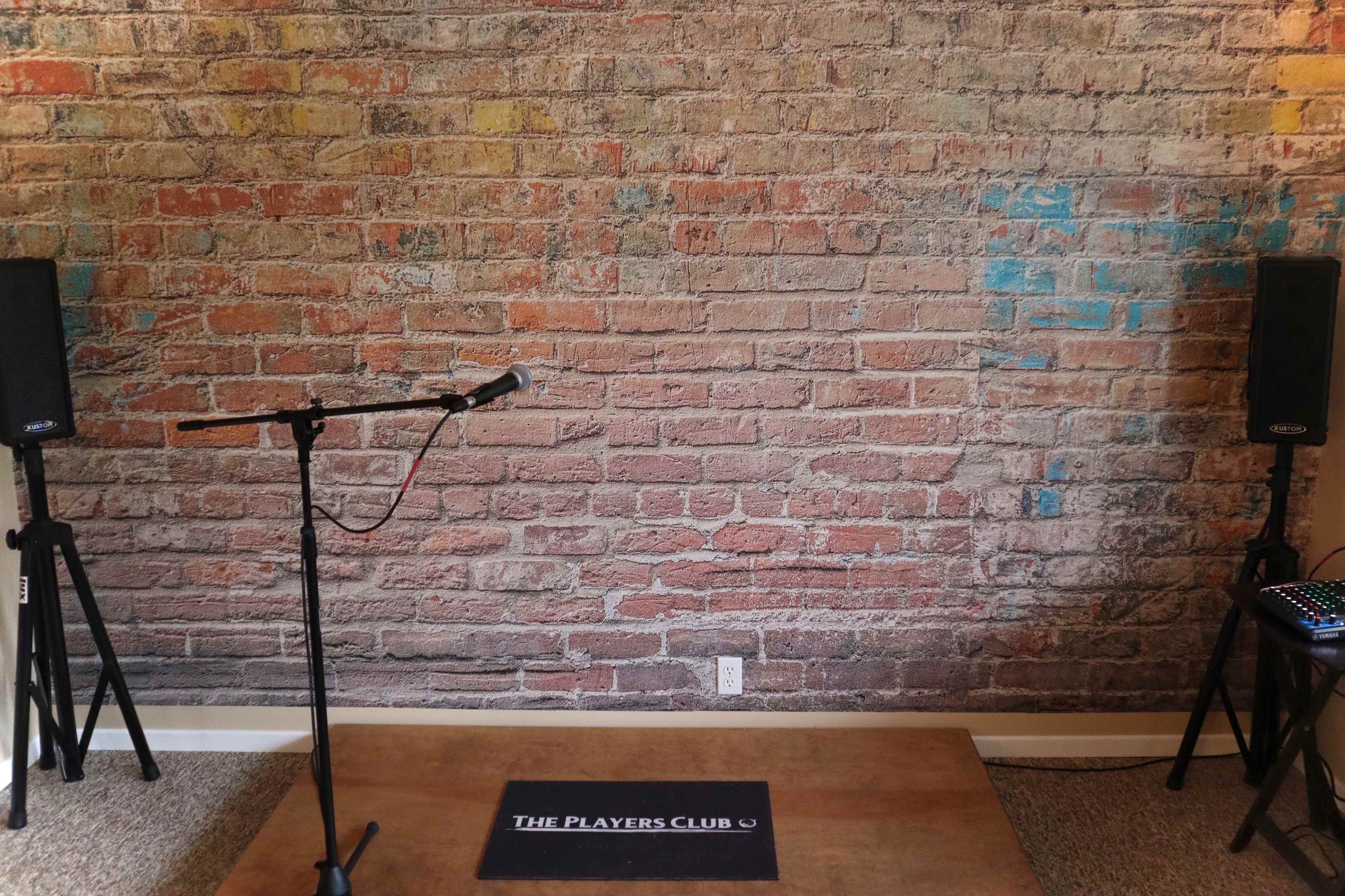 A small performance stage with a microphone stand and two speakers set against a brick wall backdrop.