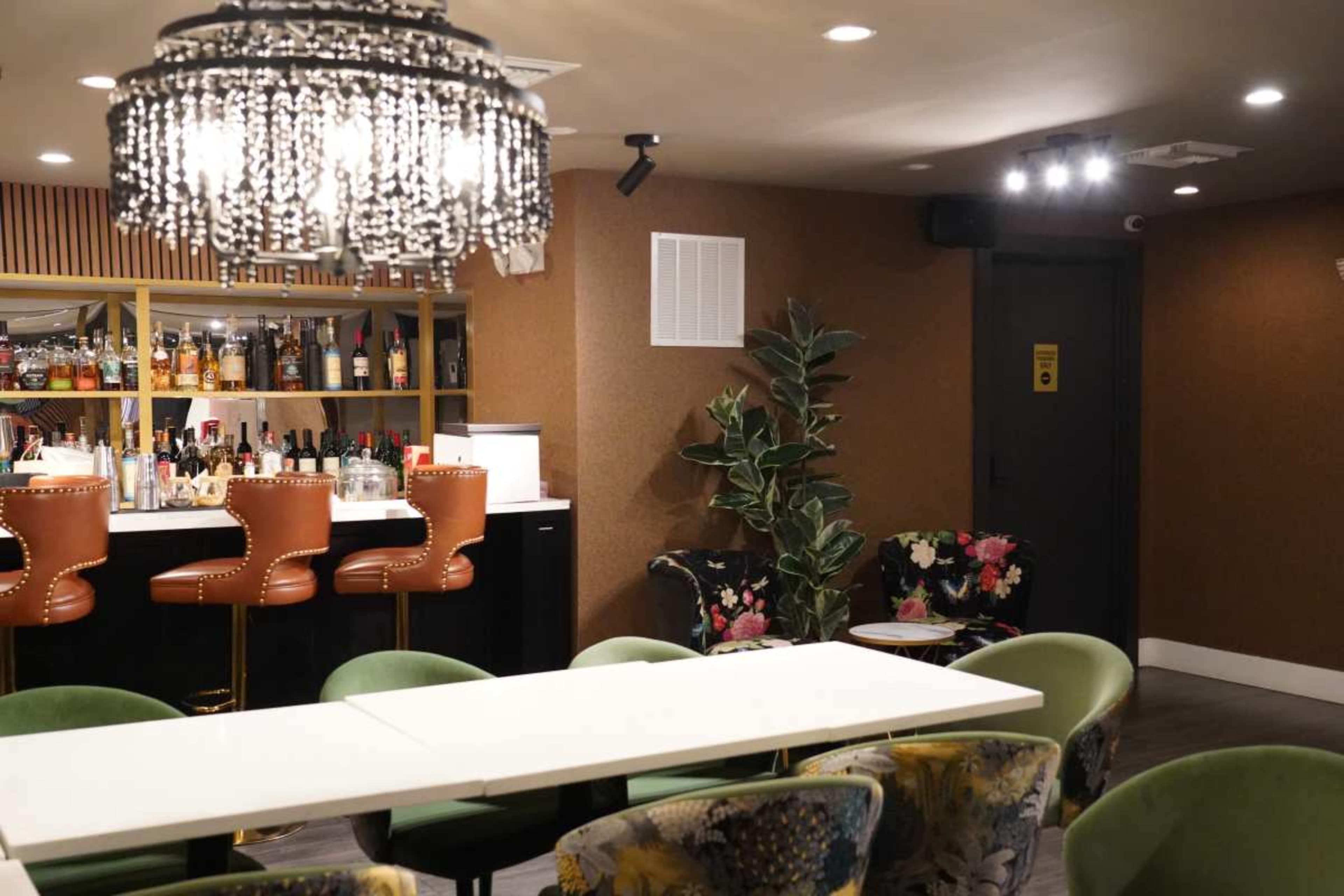 The image shows a modern lounge area with a bar featuring high-backed chairs, a variety of drinks on shelves, and decorative plants, alongside circular tables and floral-patterned seating.