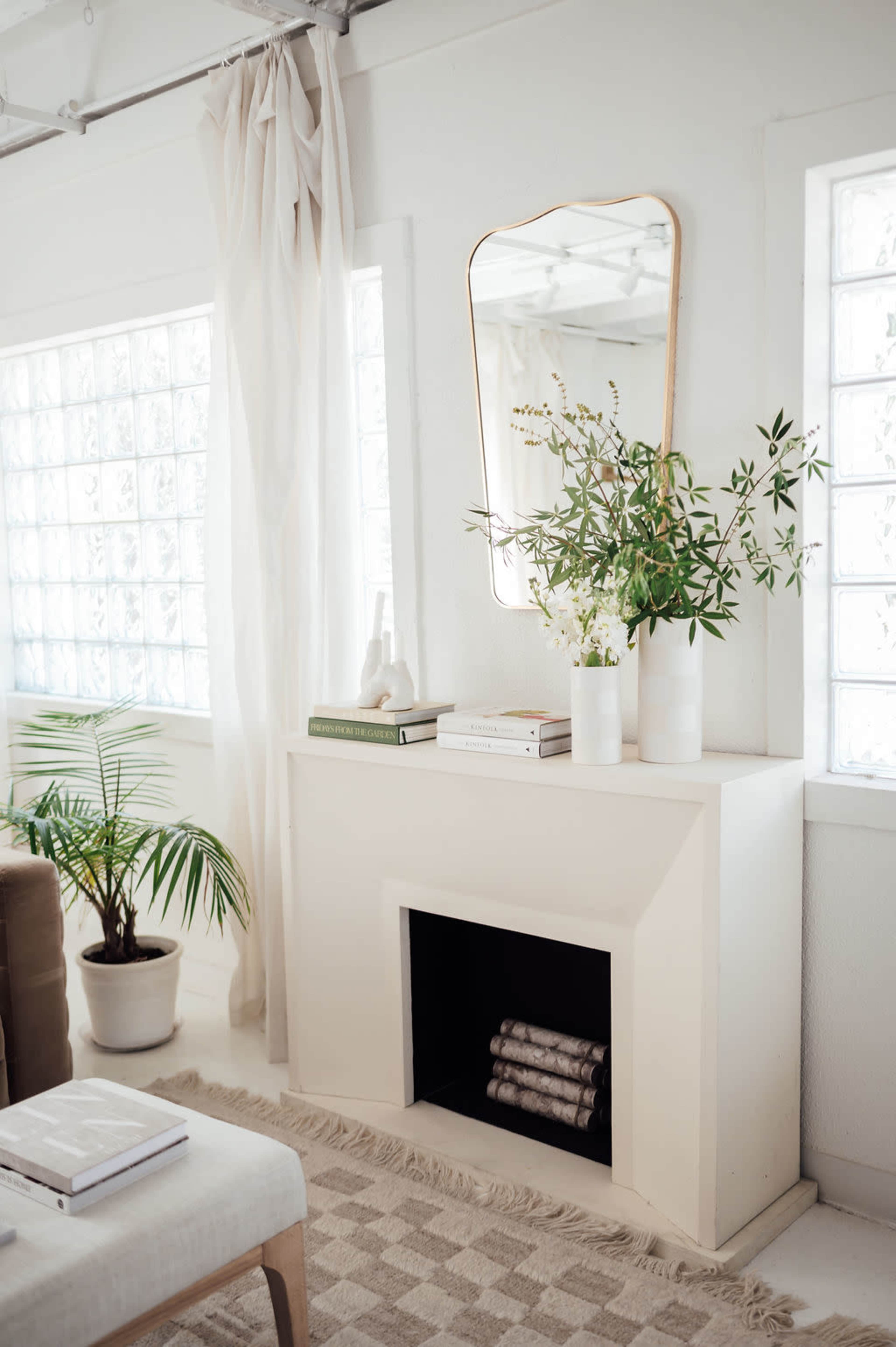 The image shows a light, minimalist living space featuring a white cabinet with a mirror above it, plants, books, and decorative vases.